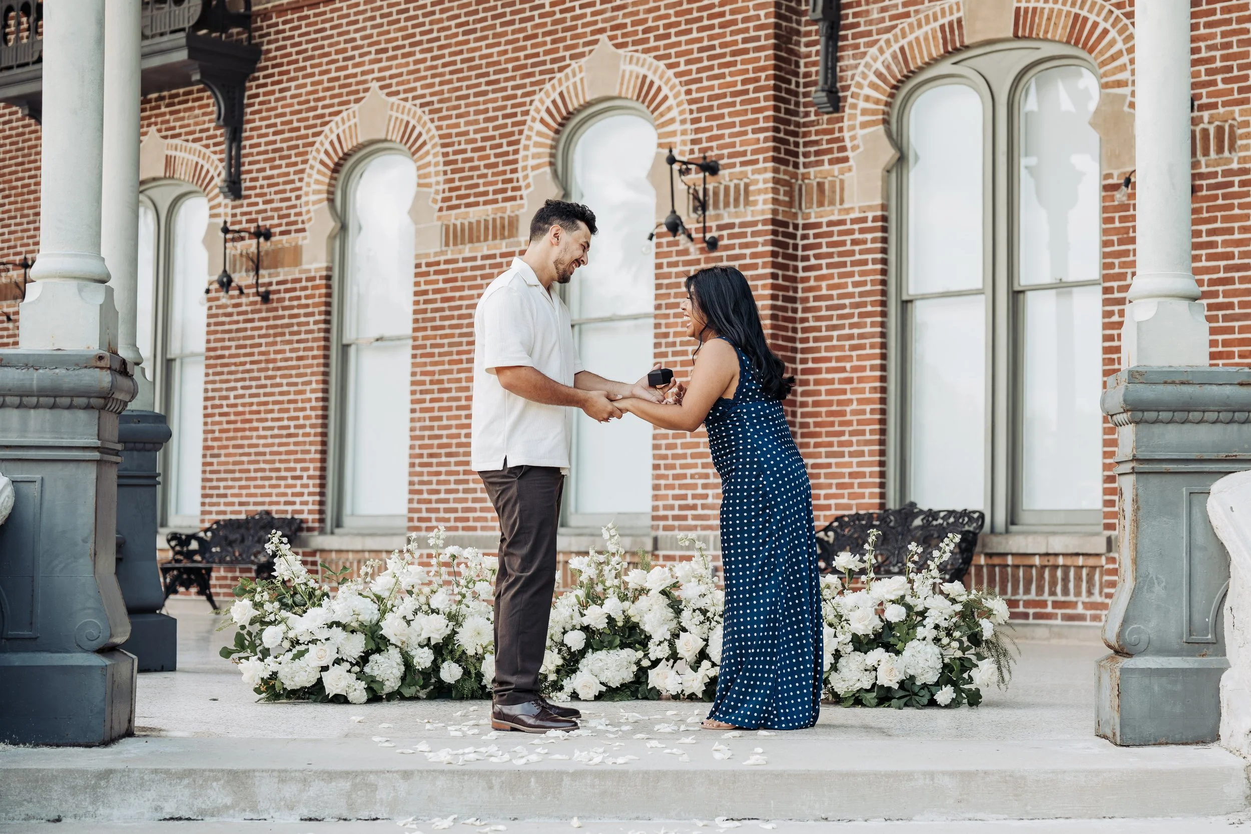 A couple exchanging rings during a wedding ceremony outside a brick building with arched windows. The man is wearing a white shirt and brown pants, and the woman is in a navy blue polka-dot dress. Flowers are arranged at their feet, and they are hold