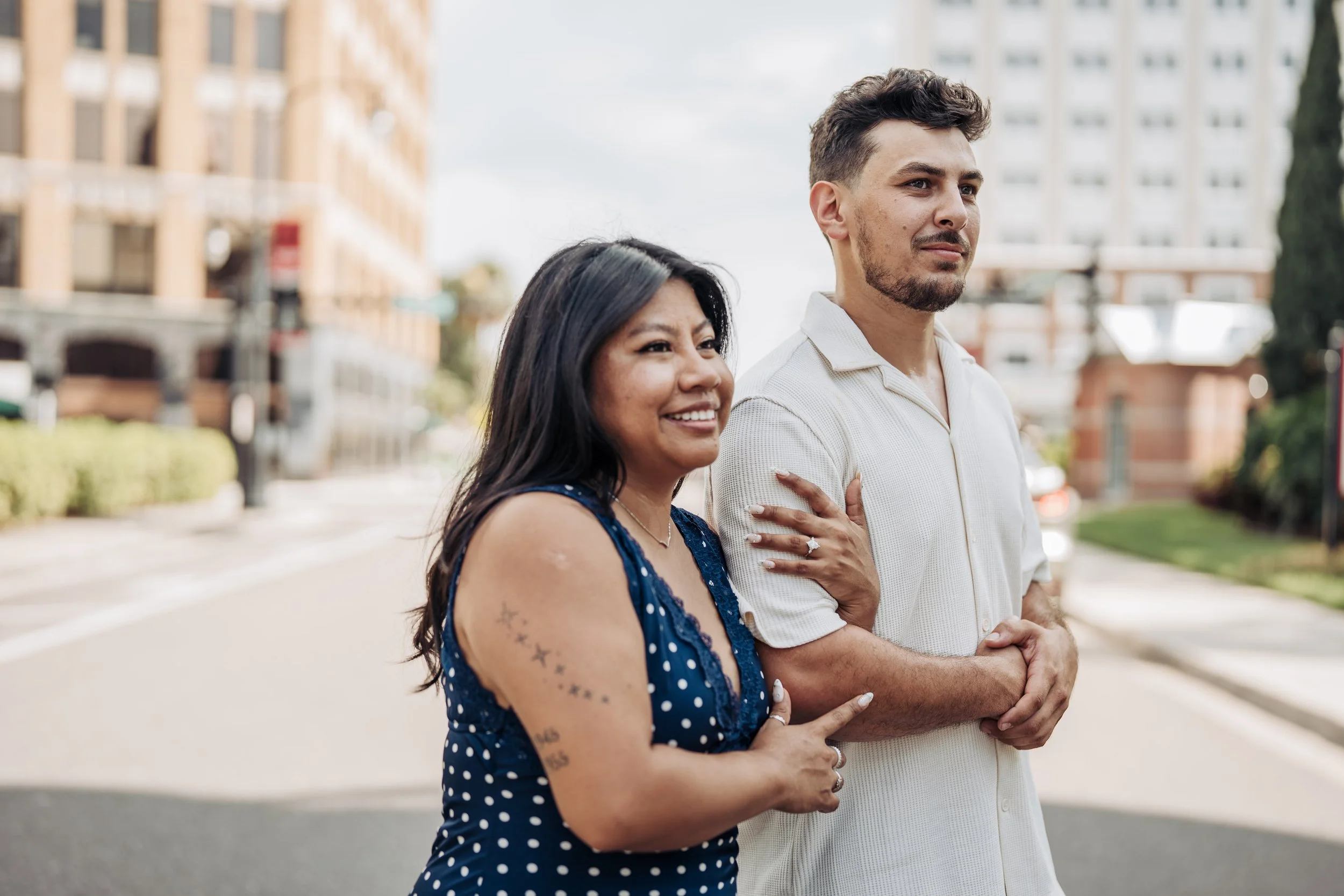 A woman and a man standing outdoors in an urban area. The woman has long black hair, tattoos on her arm, and is wearing a blue polka dot dress. The man has short dark hair, facial hair, and is wearing a white collared shirt. They are smiling and look