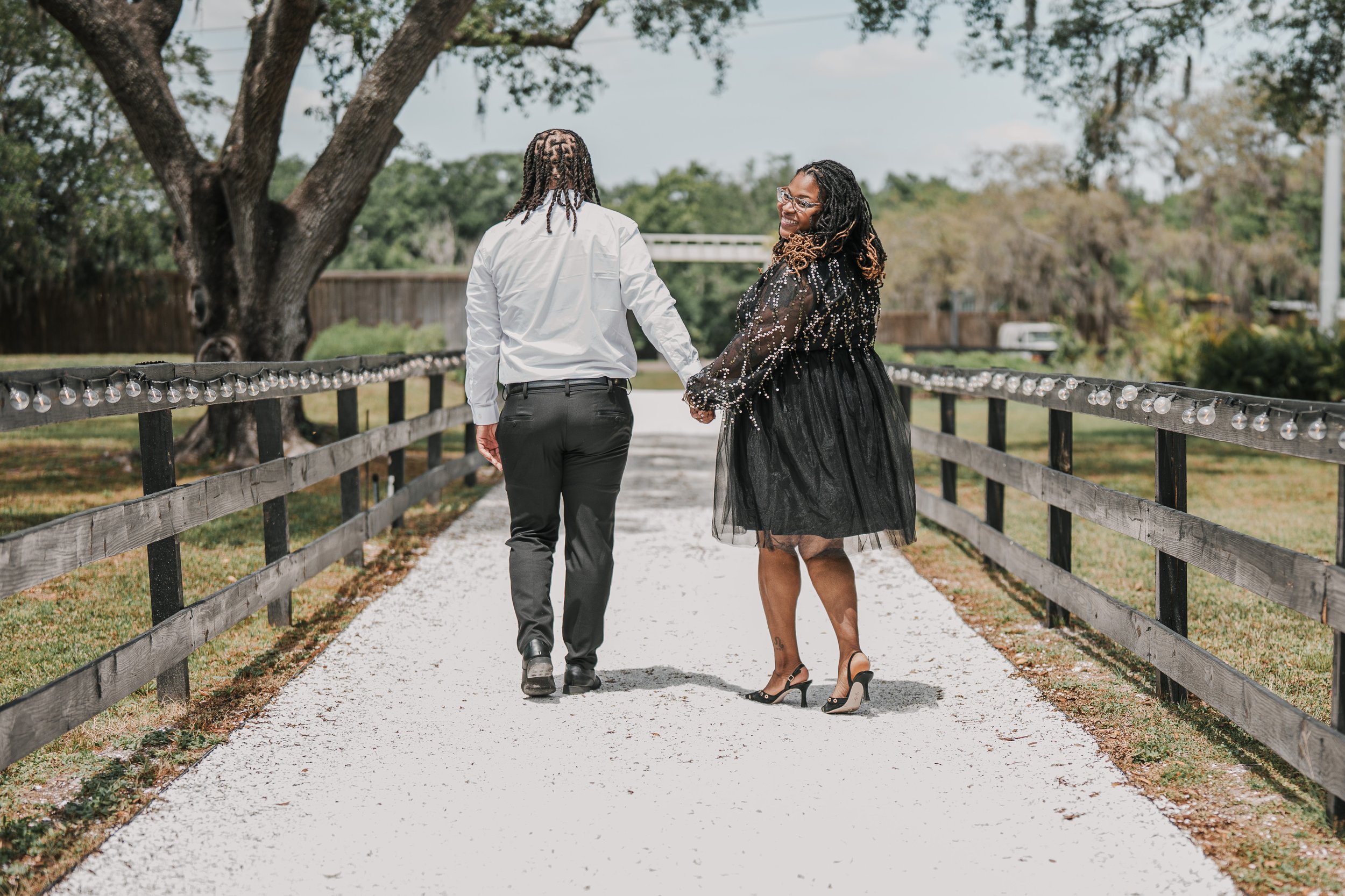 A man and woman holding hands and walking on a wooden bridge in a park. The man is dressed in a white shirt and dark pants, while the woman wears a black, sparkly dress and high heels. There are trees and a cloudy sky in the background.