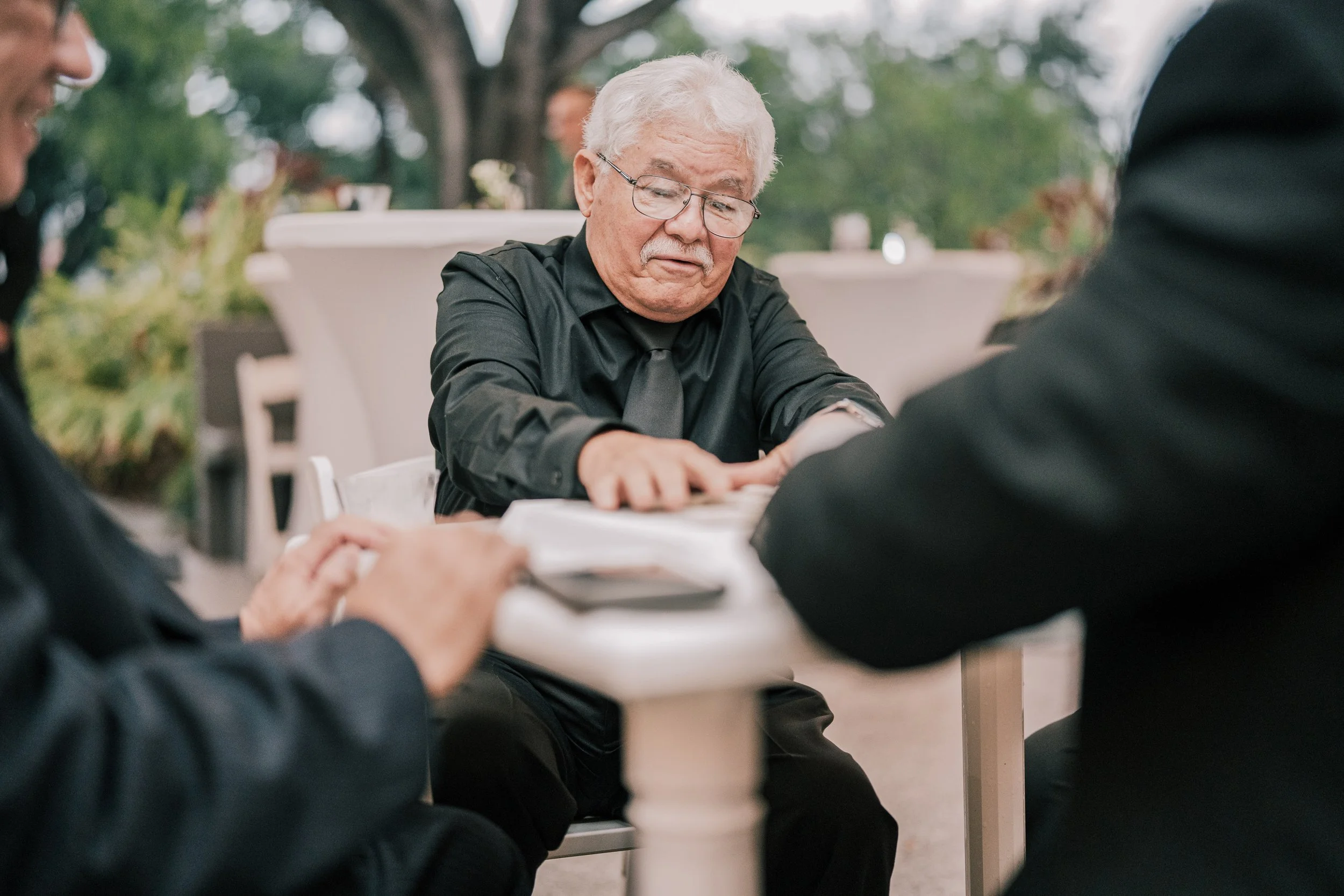 Older man with gray hair and glasses, wearing a black shirt and tie, sitting at a table with other people, outdoors, with trees in the background.
