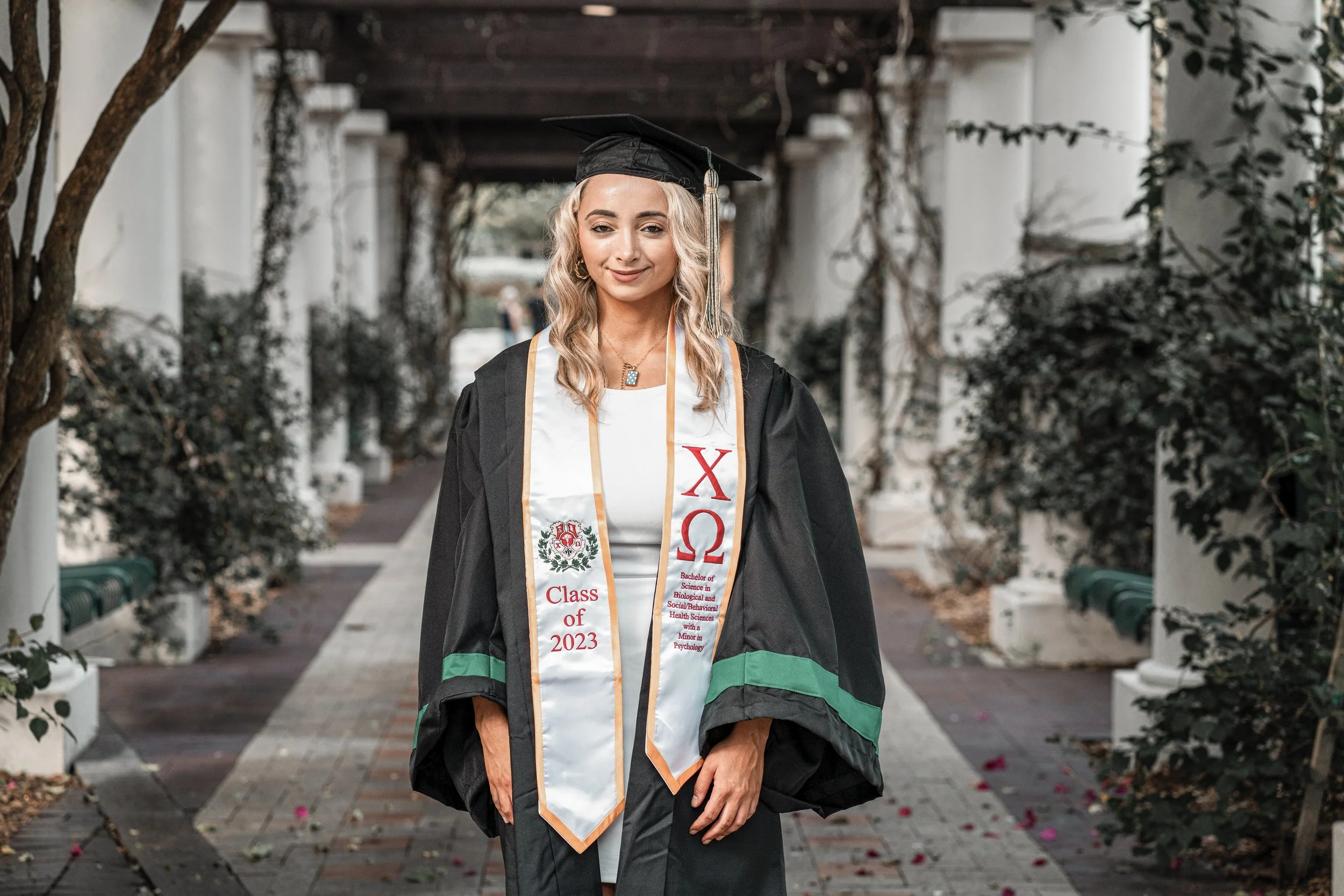 A young woman in a graduation cap and gown standing outdoors in a walkway lined with white pillars and greenery, holding a sash that reads 'Class of 2023' from Harvard.
