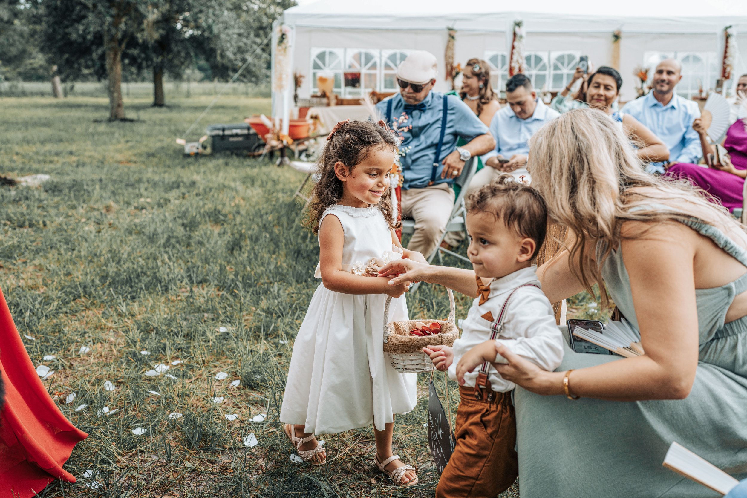 A young girl in a white dress exchanging a basket of red apples with a young boy at an outdoor event, with seated adults and a decorated tent in the background.
