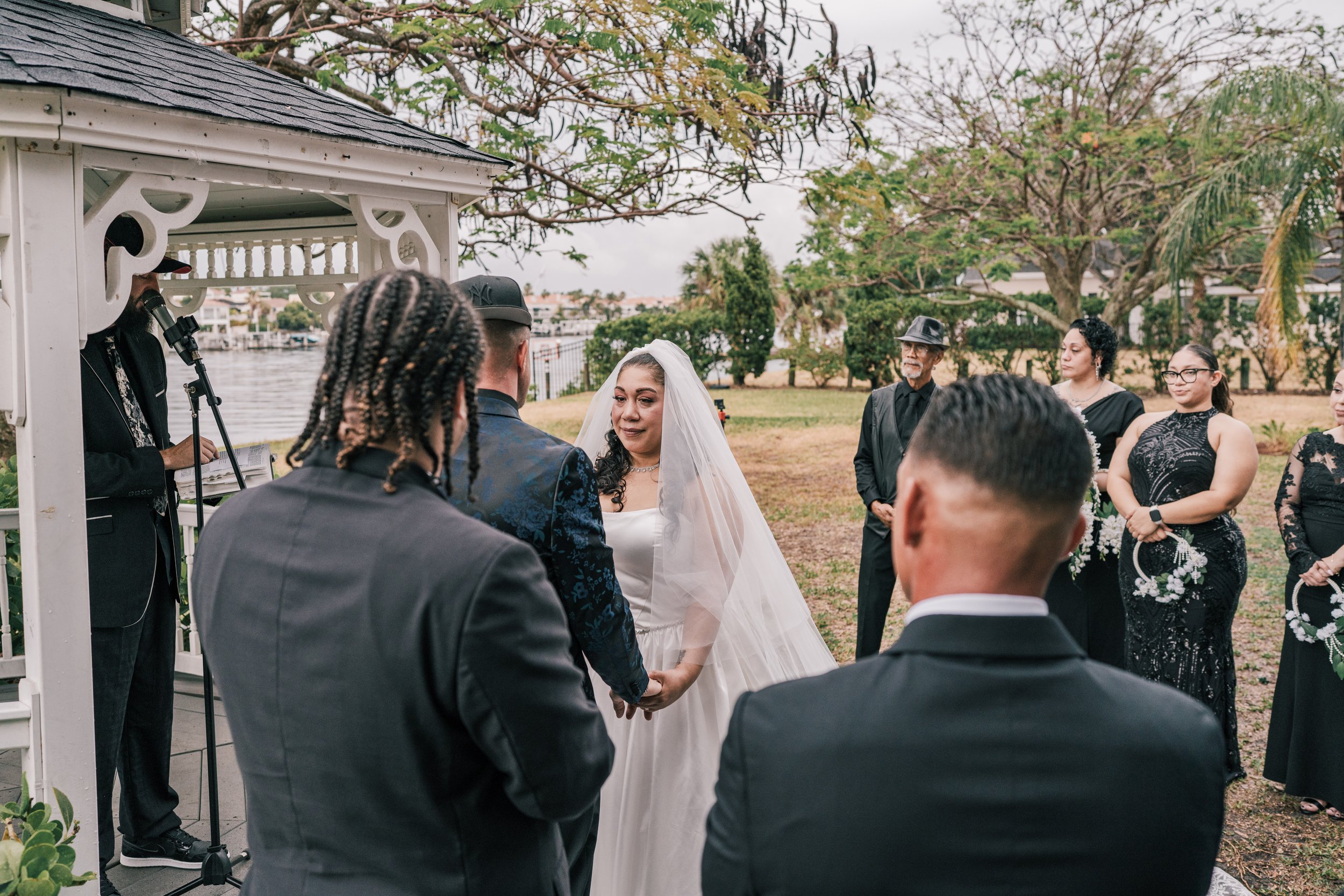 A wedding ceremony outdoors near a body of water, featuring a bride in a white wedding dress and veil, a groom in a dark suit, and guests in formal attire, with a officiant speaking into a microphone.
