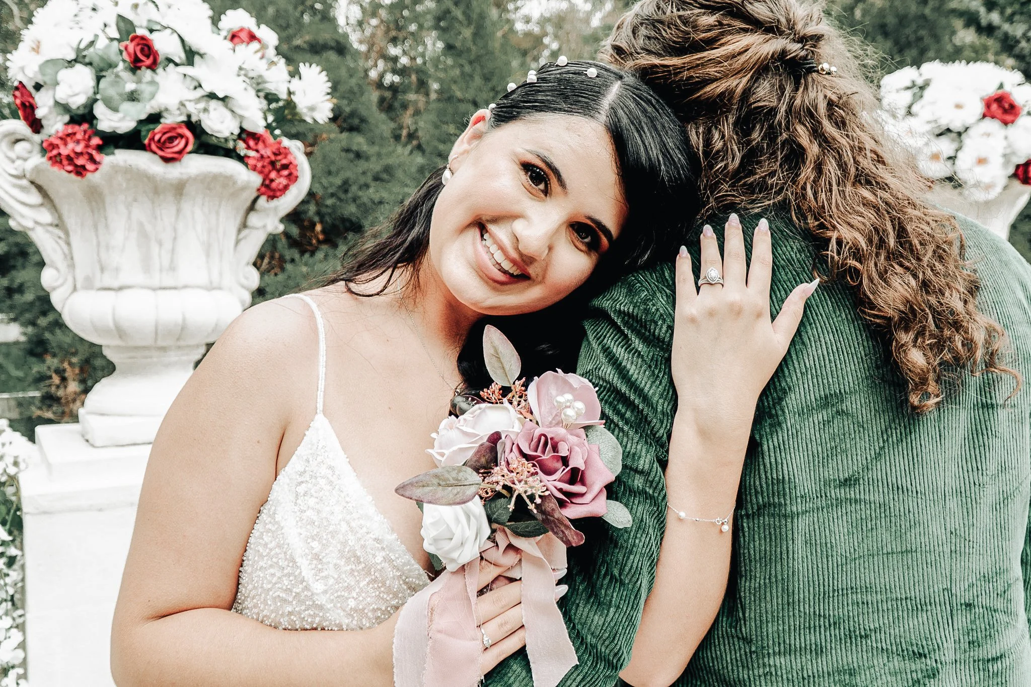 A woman in a white dress holding a bouquet of flowers, hugging another person outside near potted flowers.