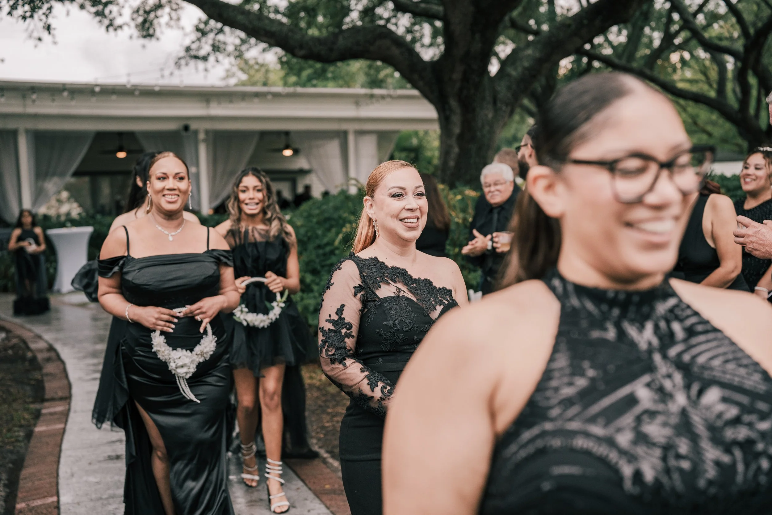 Group of women dressed in elegant black dresses standing outdoors under a large tree at an event, smiling and chatting.