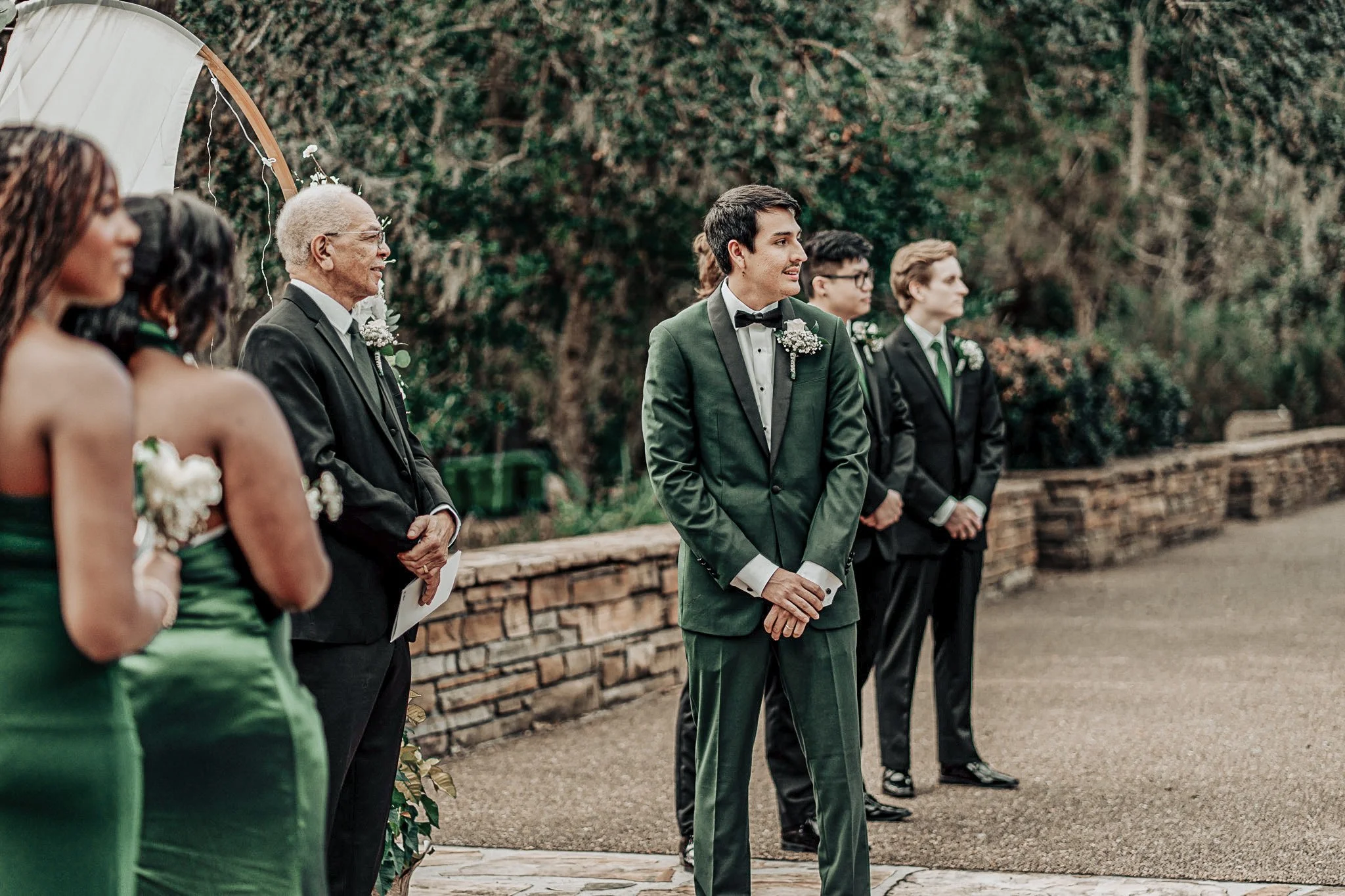 A wedding ceremony outdoors with wedding party members in formal attire, standing in a line on a gravel path, with trees and a wooden fence in the background.
