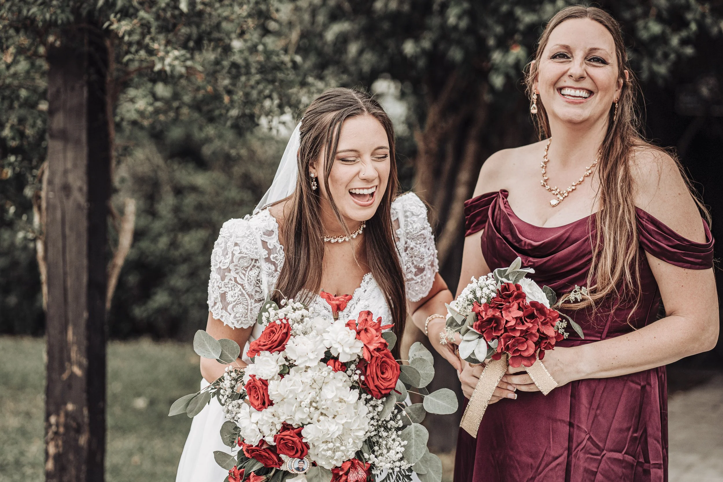 Two women, one in a white lace wedding dress and the other in a burgundy gown, are smiling and holding bouquets of red and white flowers in an outdoor setting with trees in the background.