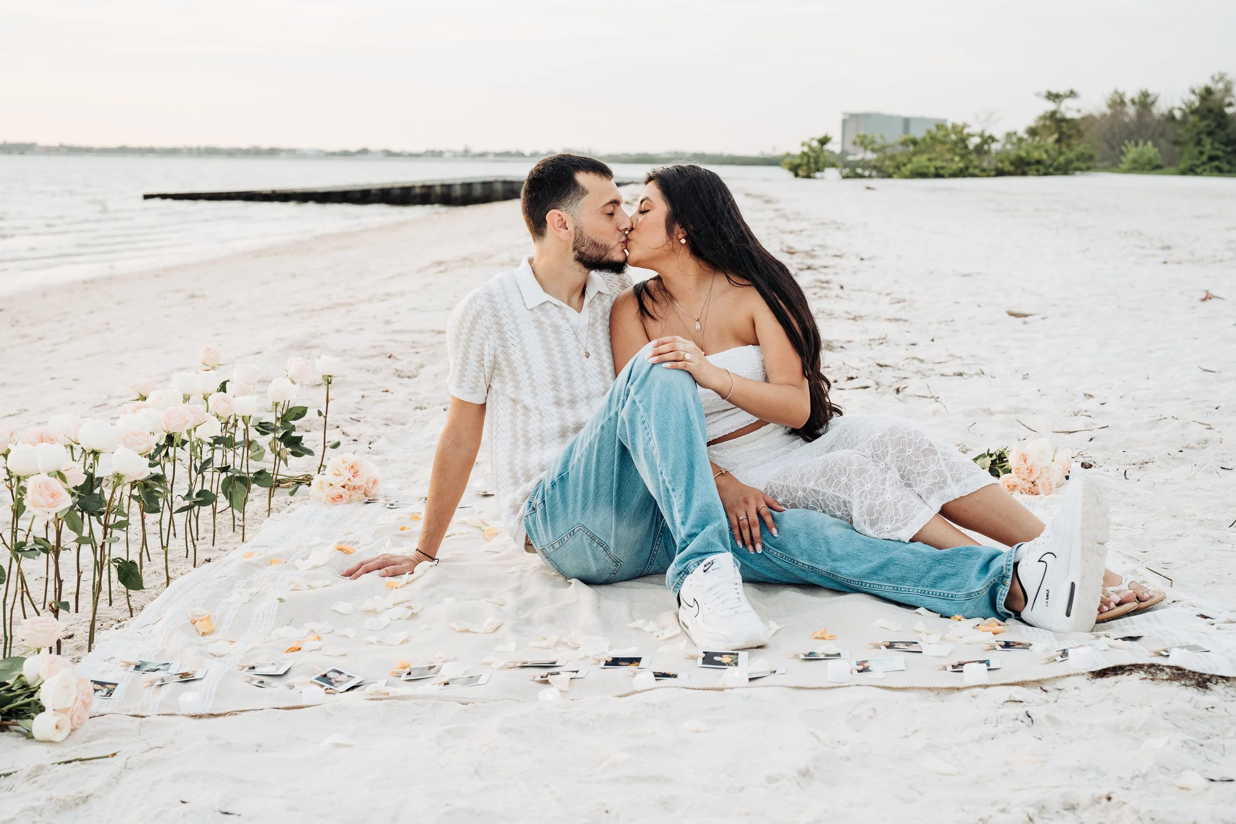 A couple kisses on a sandy beach during a romantic picnic setup with roses and photographs scattered on a blanket.