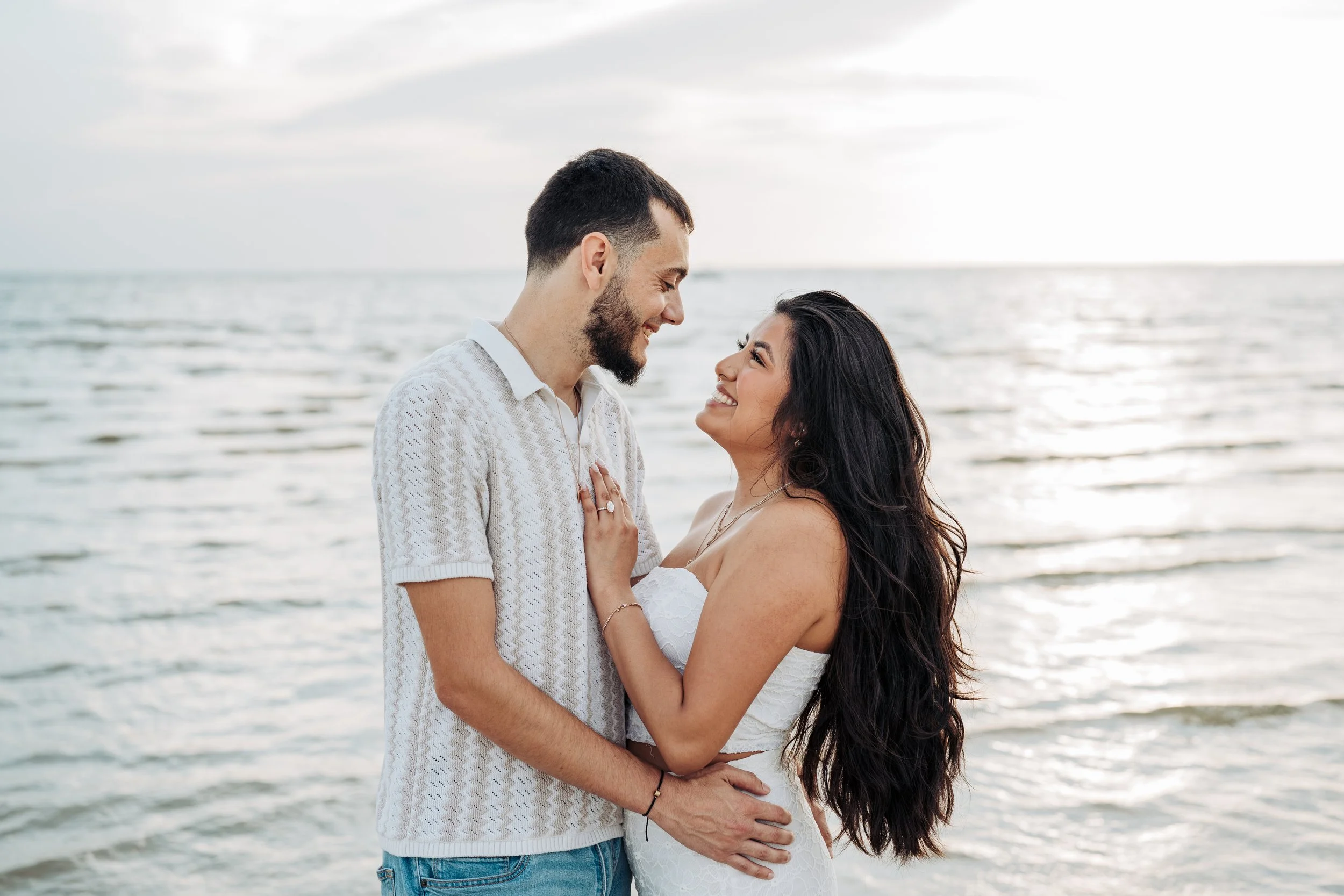 A happy couple standing close together by the water at the beach, smiling and looking into each other's eyes.