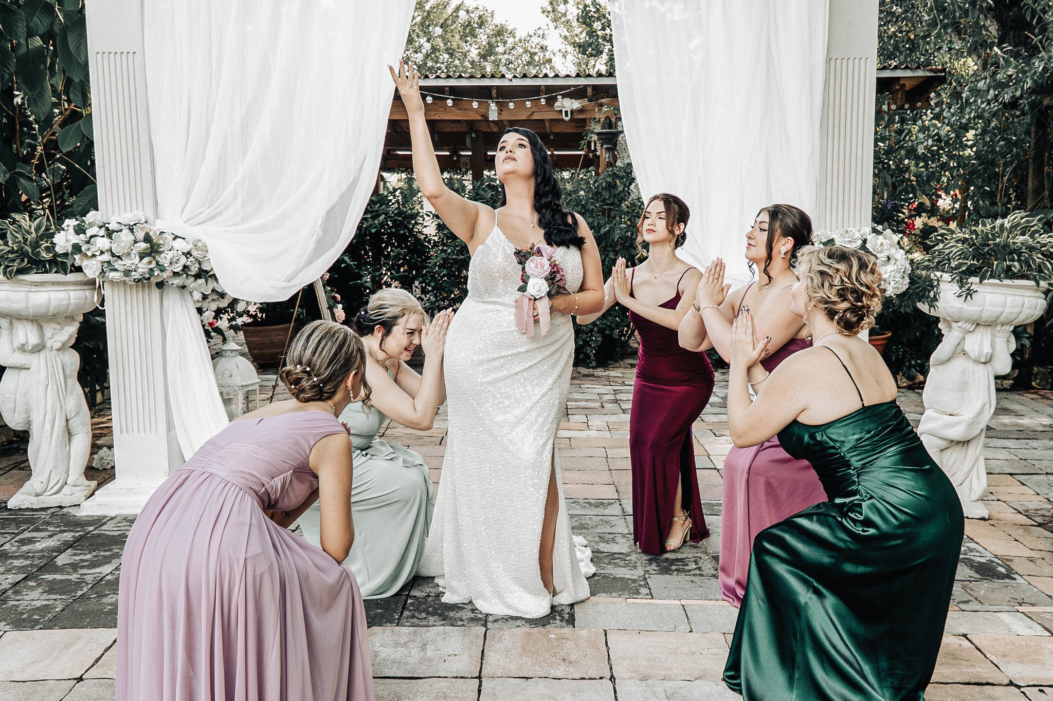 A bride in a white wedding dress holding a bouquet is standing with her eyes closed as she is being blessed or prayed over by several women dressed in formal gowns in an outdoor setting with white drapery and greenery.