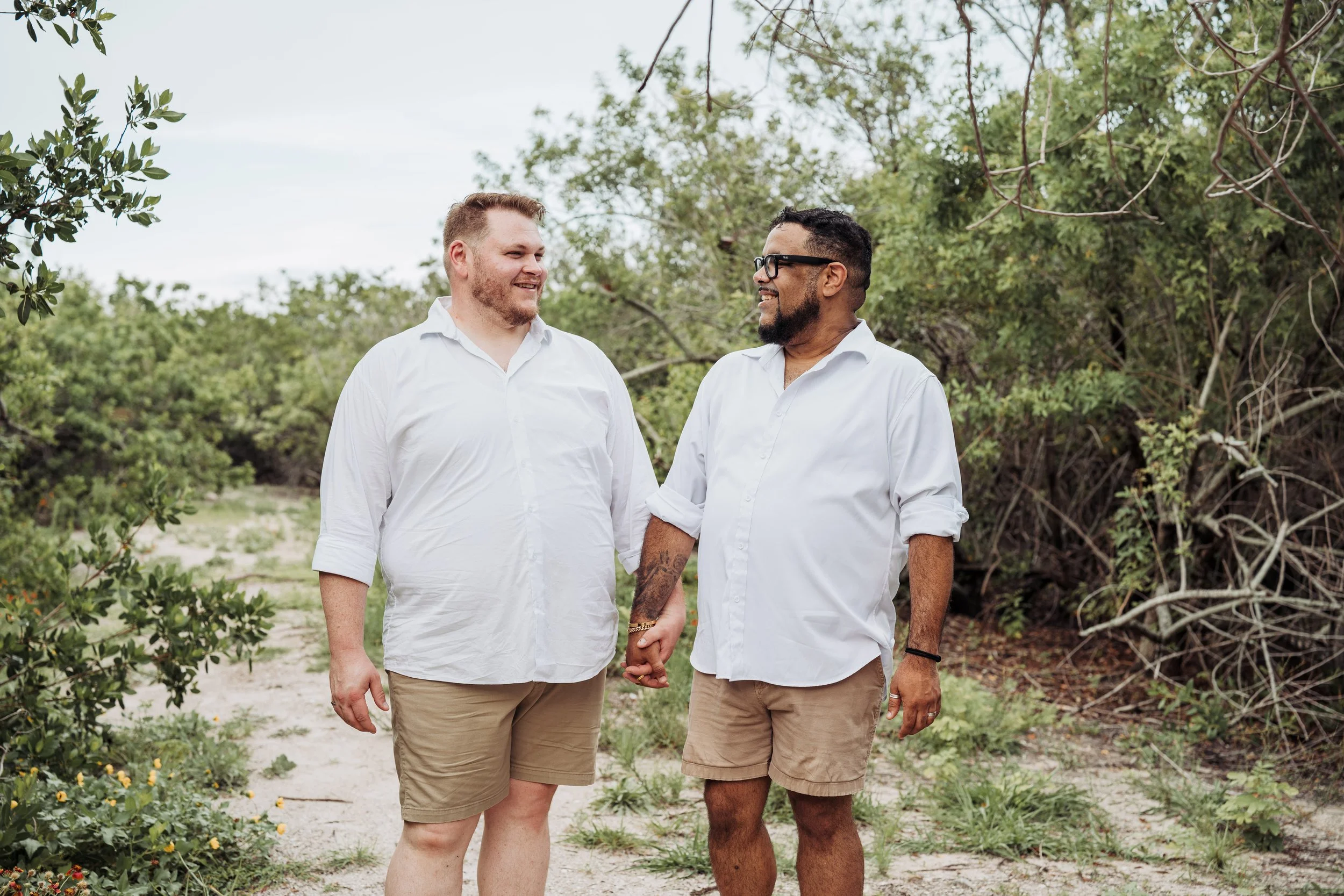 Two men dressed in white shirts and shorts are holding hands and smiling at each other while standing outdoors in a green, wooded area.