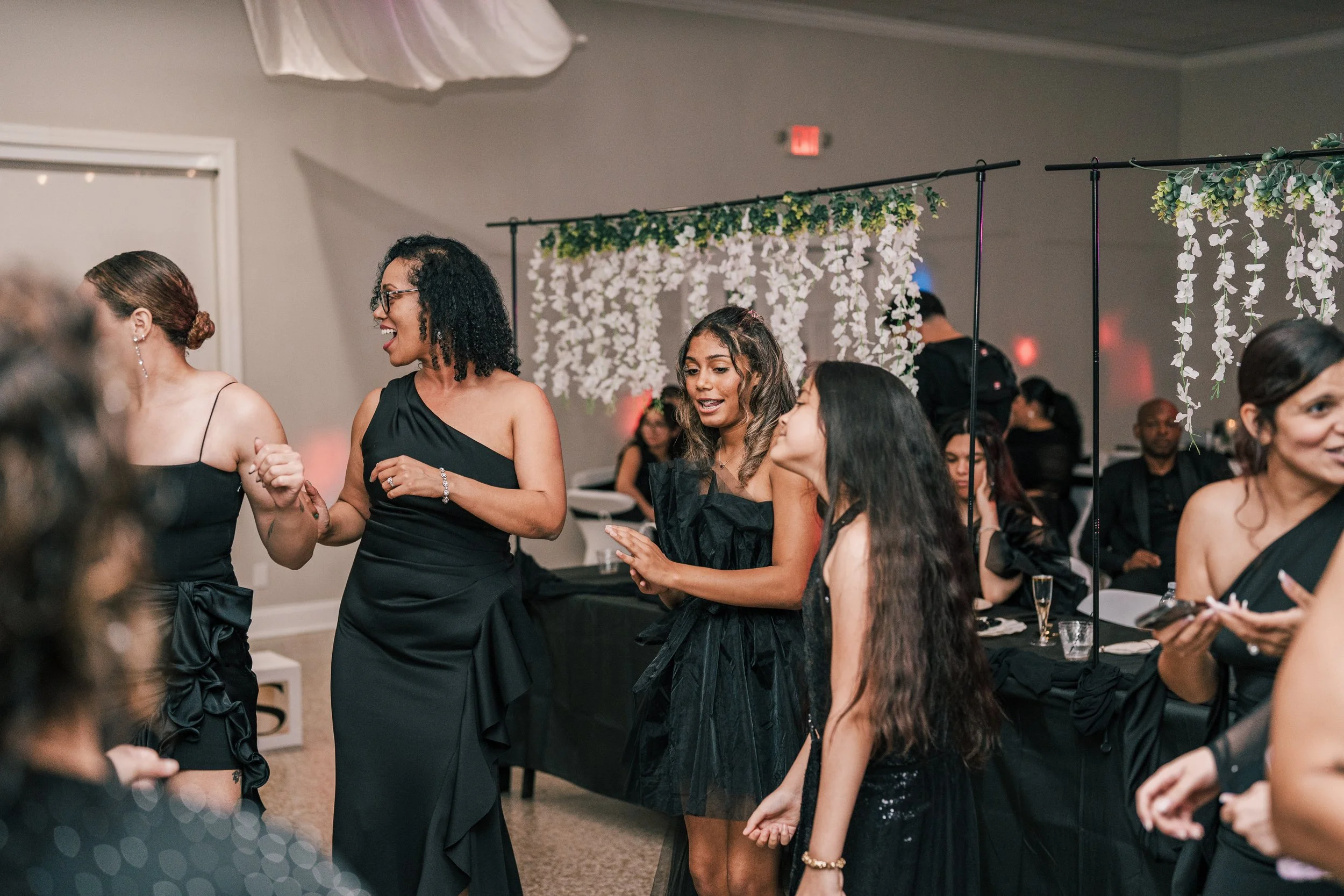 Women dancing and socializing at an event with floral decorations in the background.