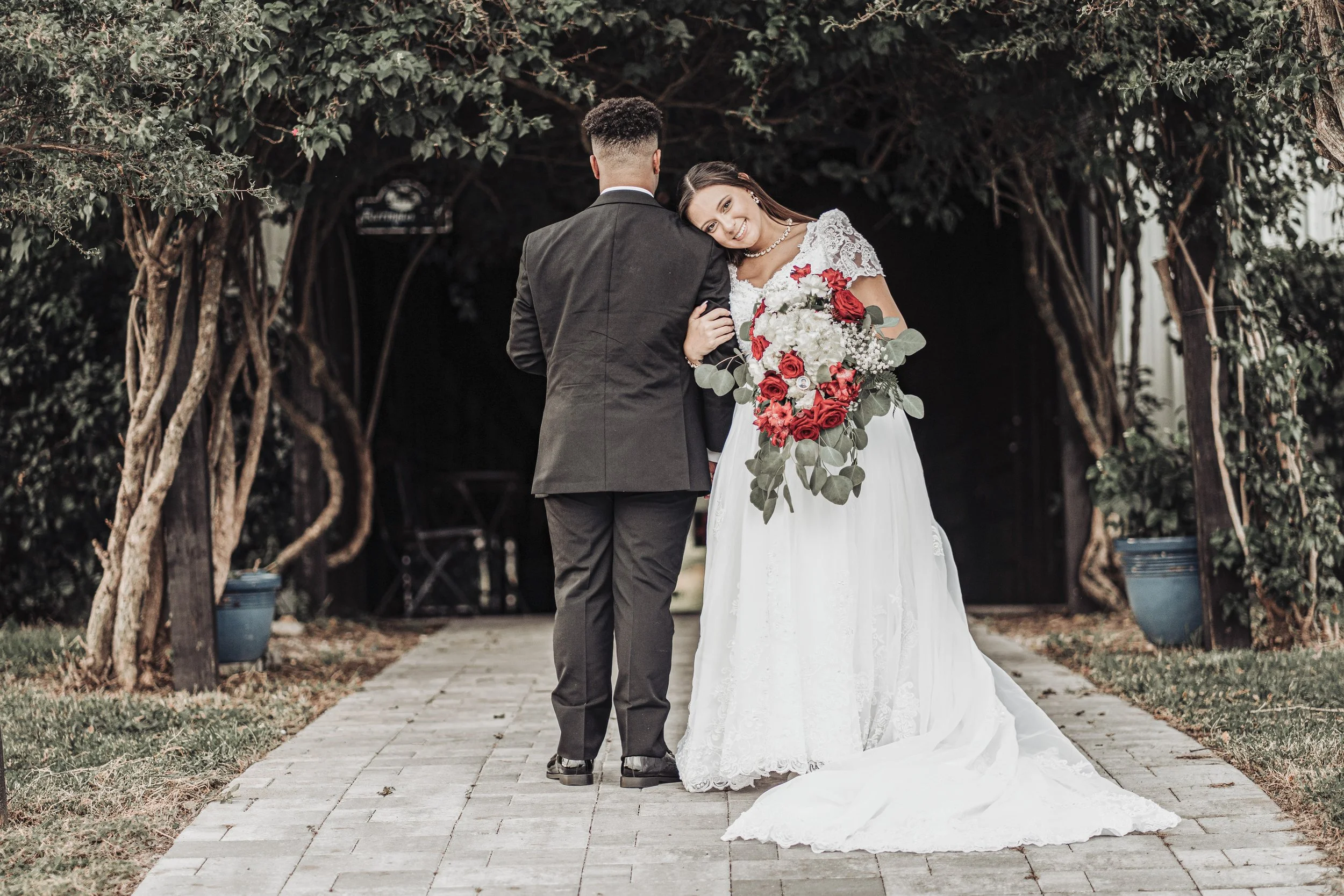A bride and groom stand outdoors in a garden with trees, with the bride holding a bouquet of red and white flowers and smiling, facing to the right while leaning her head on the groom's shoulder, who is dressed in a black suit with his back to the ca