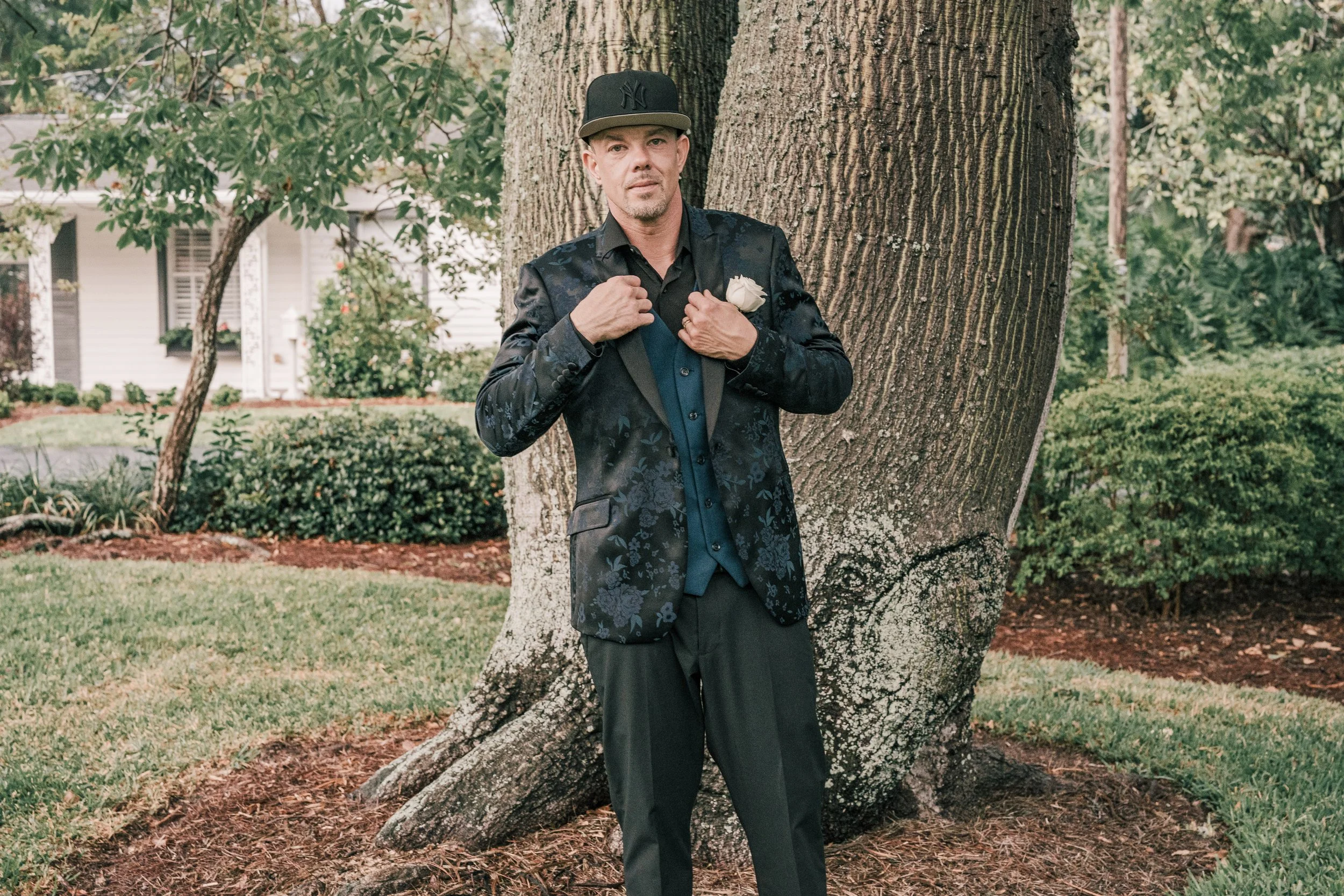 A man in a dark, floral patterned suit with a white boutonniere on his lapel stands outdoors, leaning against a large tree with his hands adjusting his shirt. He is wearing a black mixed with dark blue shirt, a matching jacket, a black and gray New Y