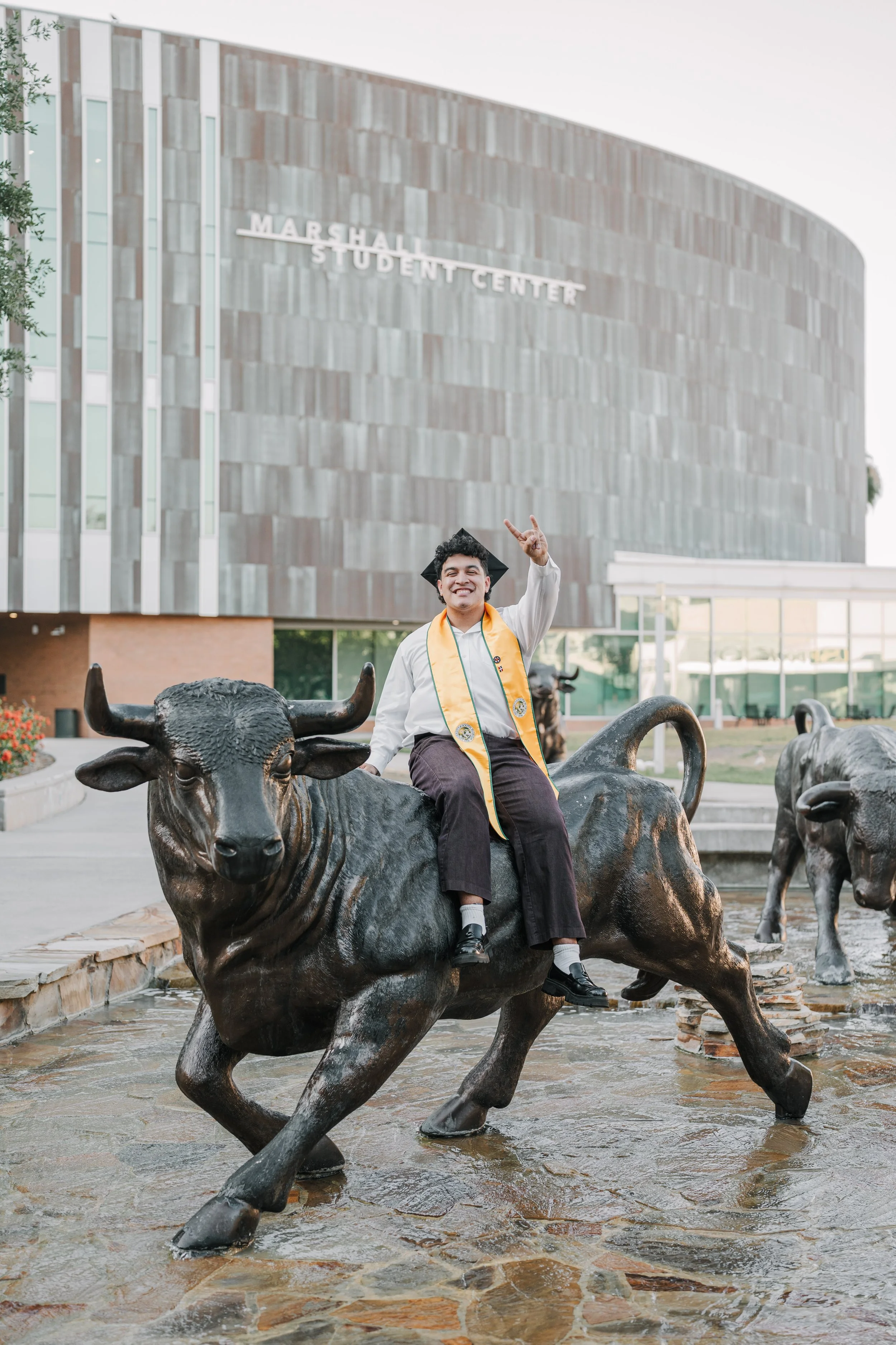 A graduate in cap and gown sitting on a bronze bull statue in front of the Marsha Student Center building, with other animal statues nearby.