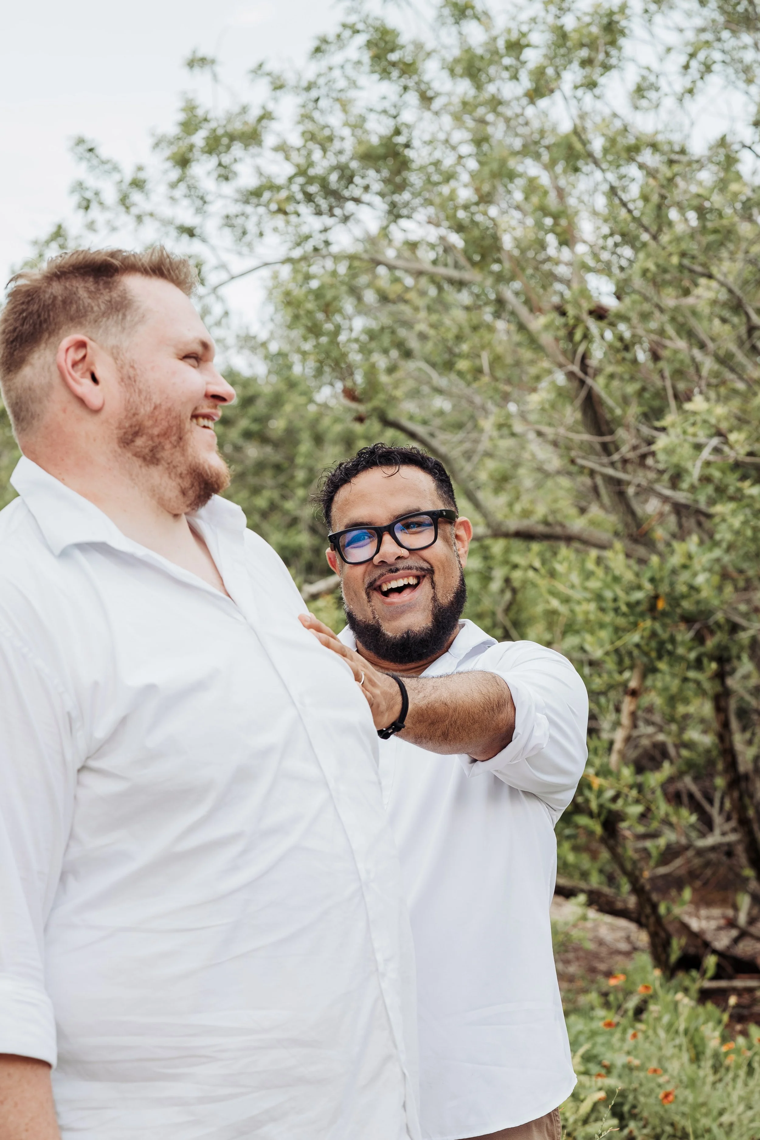 Two men smiling and laughing outdoors, one man has light skin and red hair, the other man has medium skin, black hair, glasses, and a beard. They are wearing white shirts and surrounded by trees and greenery.