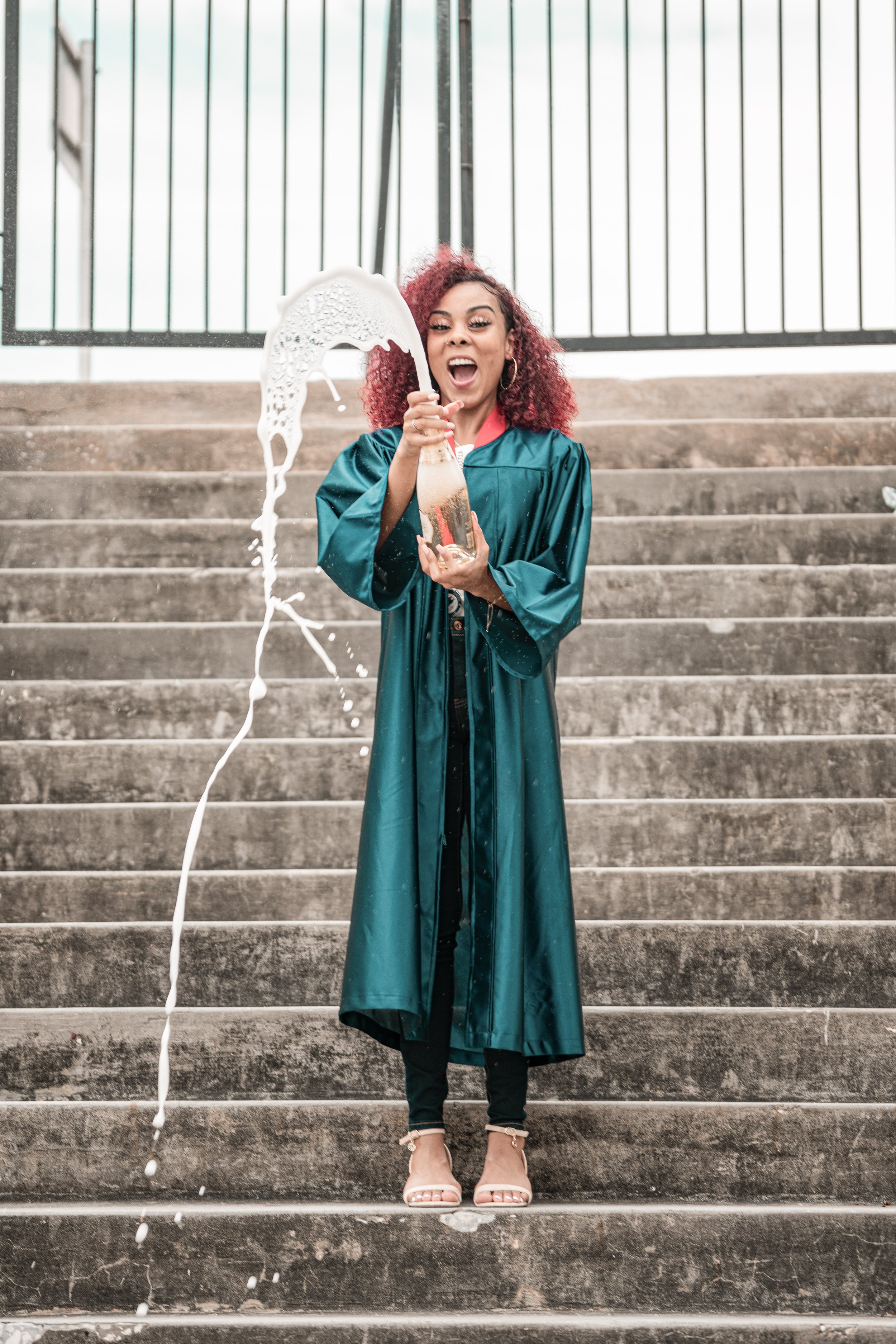 A young woman in a graduation gown standing on a stairway, holding a bottle with foam spilling out, and wearing high heels.
