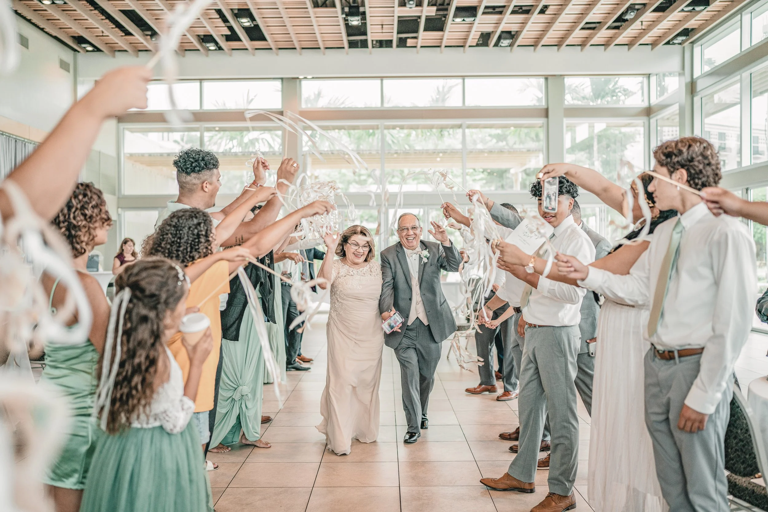 Wedding celebration with a bride and groom walking through a tunnel of people holding streamers in a bright indoor venue with large windows.
