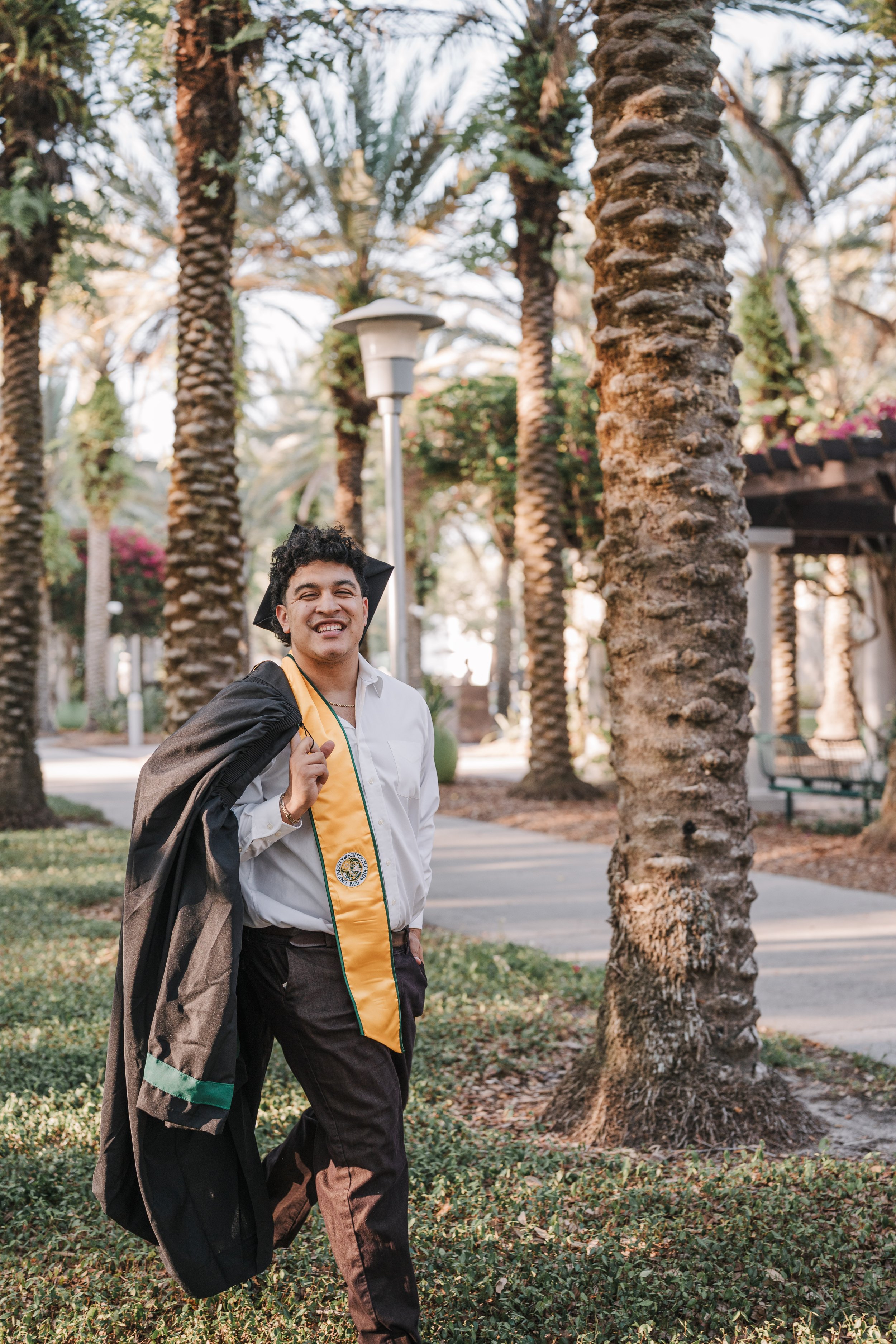 A young man in a white shirt and dark pants is smiling and wearing a graduation stole, carrying a graduation gown over his shoulder, walking in a park with palm trees and a lamppost in the background.