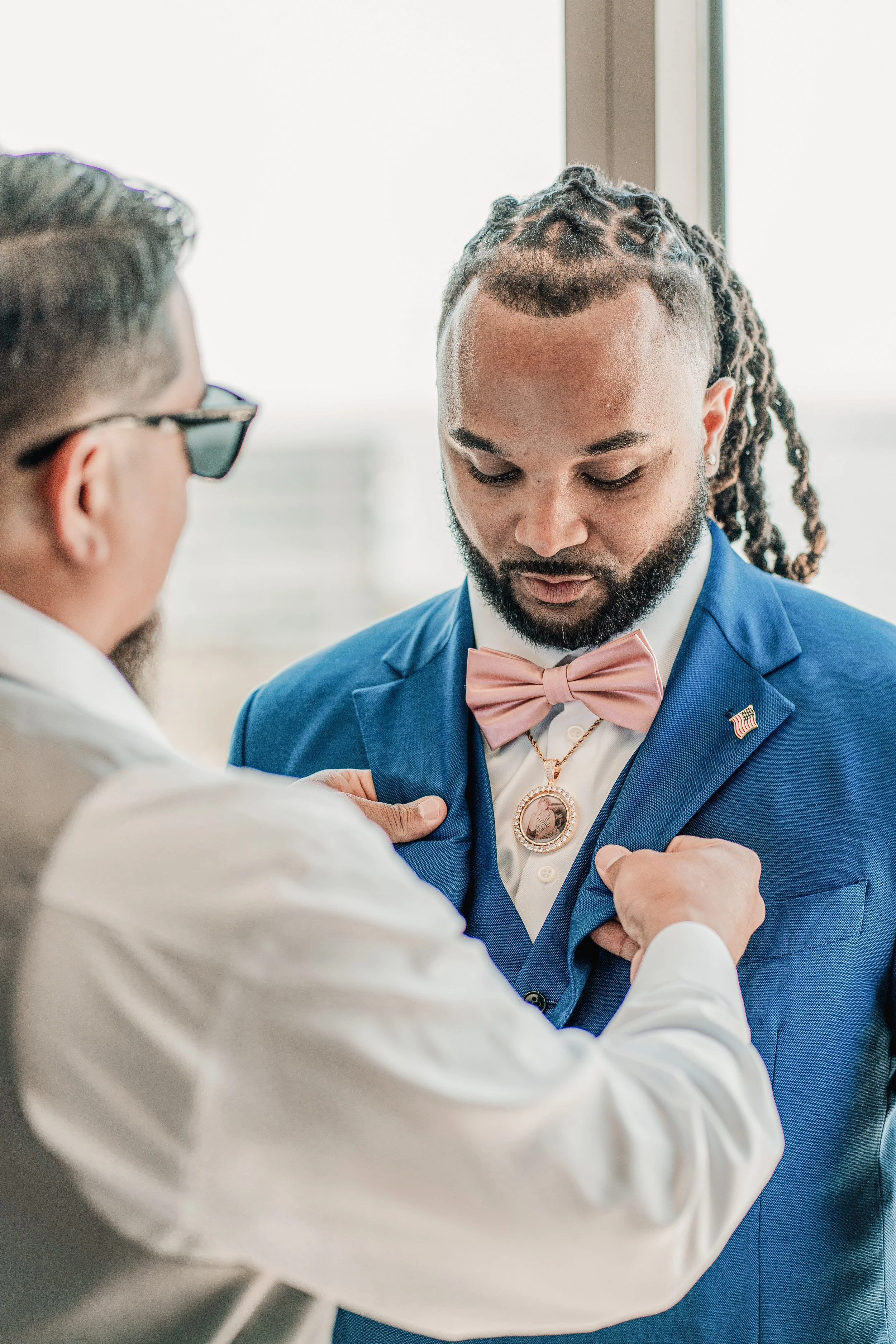 A man in a blue suit with a pink bow tie and an American flag pin is being pinned with a boutonniere by another man in a white shirt during a formal ceremony or event.