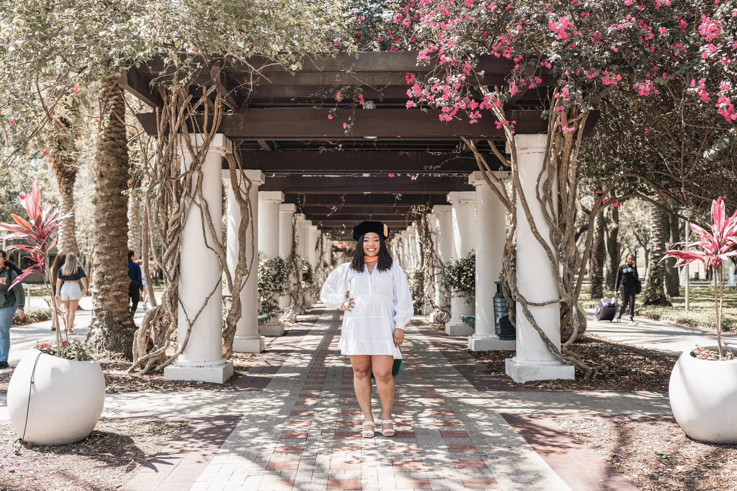A woman dressed in a white graduation gown and cap, smiling and standing outdoors under a pergola decorated with pink flowers. There are other people in the background walking and carrying luggage.