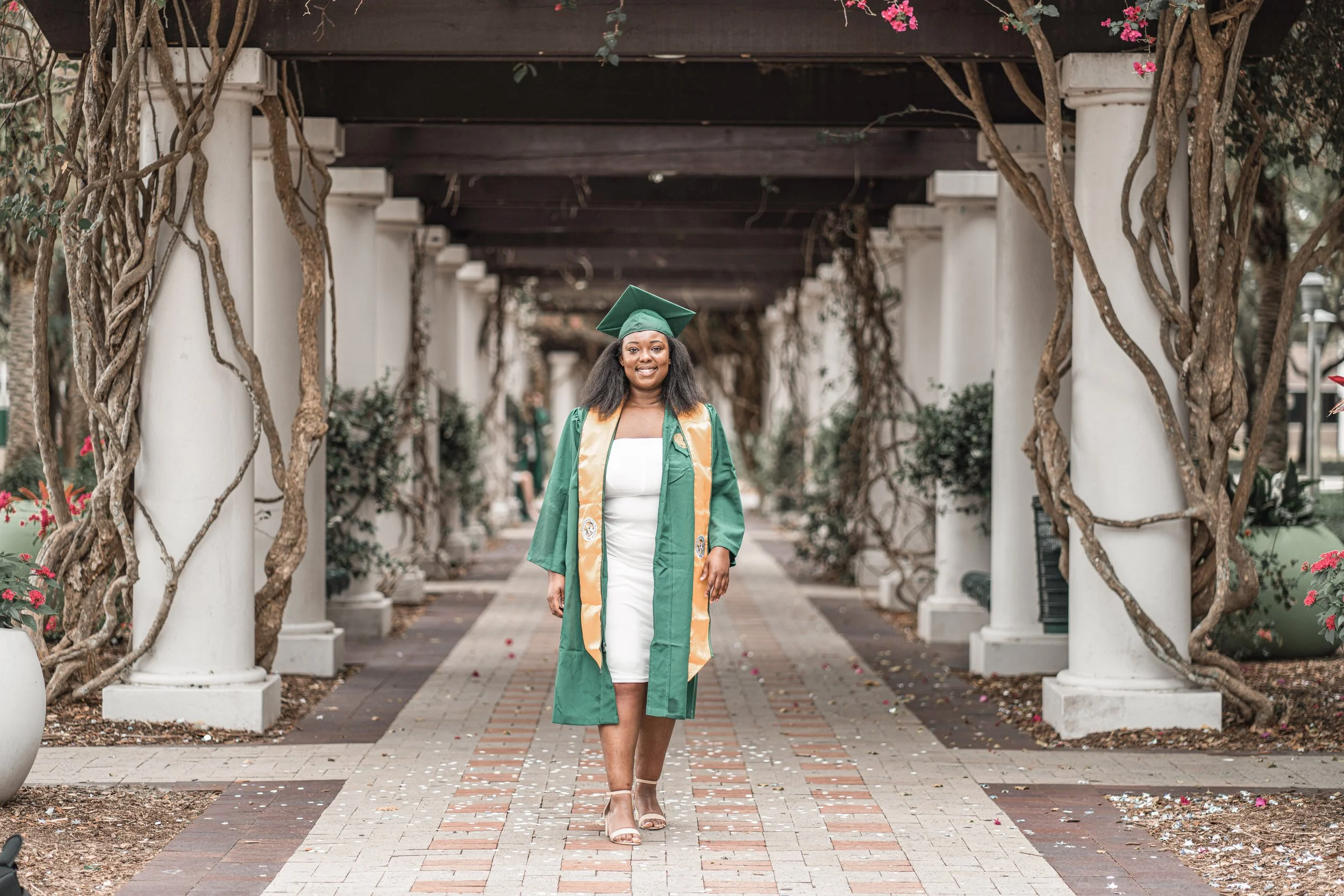 A woman wearing a green graduation cap and gown, standing on a brick-paved pathway under a pergola adorned with pink flowers and twisted tree branches, smiling at the camera.