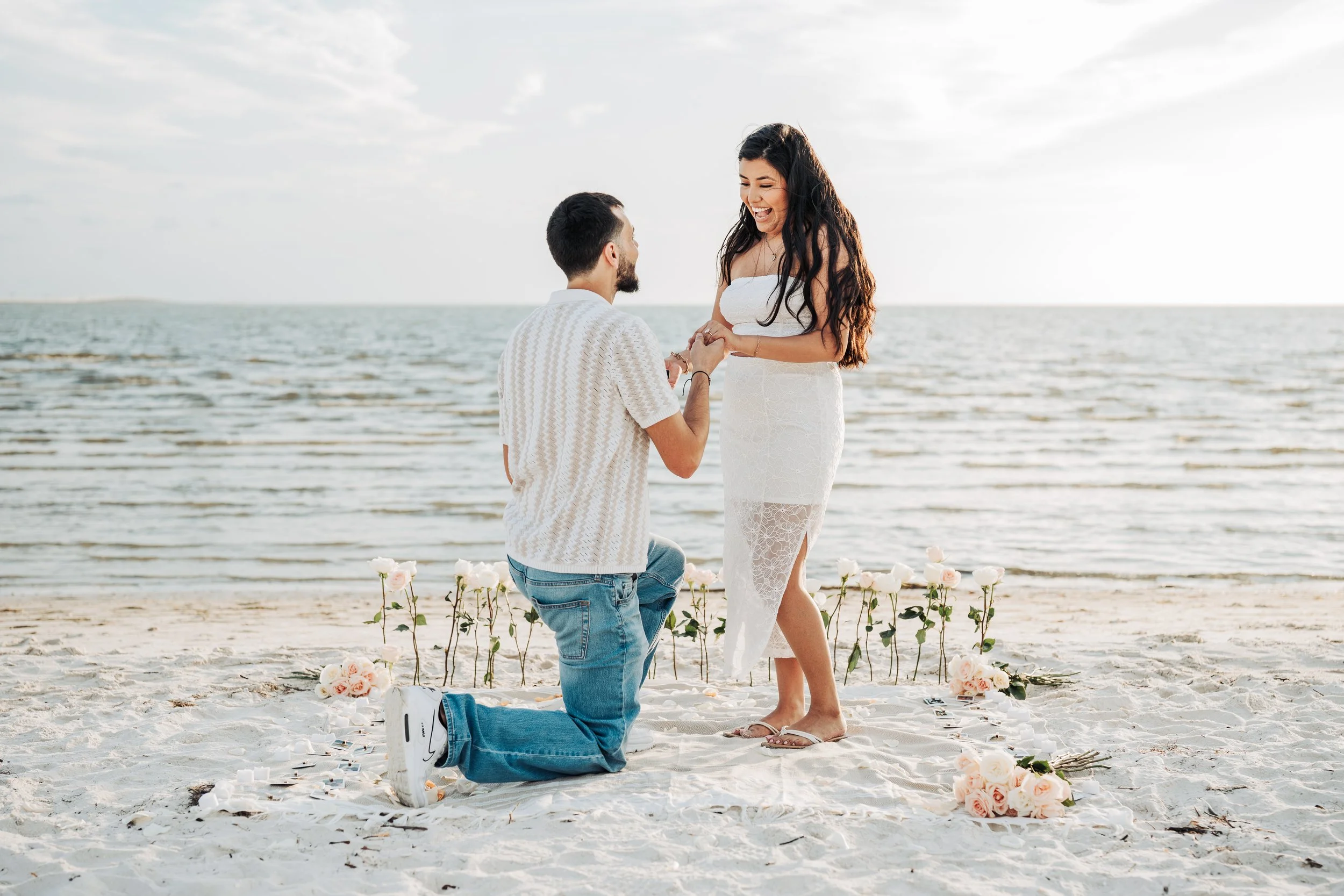 A couple getting engaged on the beach. The man is kneeling on one knee, holding the woman's hands, in front of floral arrangements and wedding decorations with the ocean in the background. The woman is smiling happily.