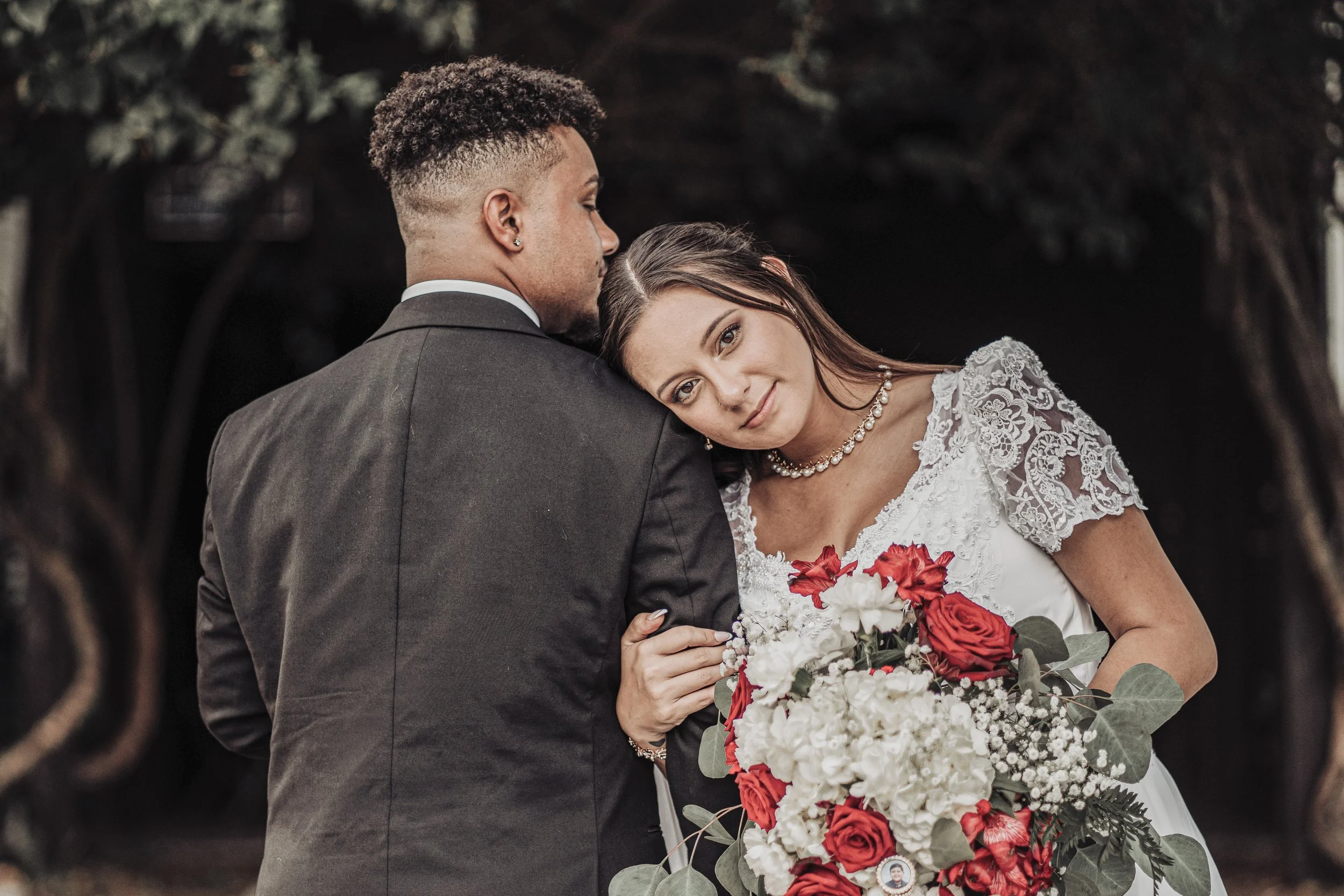 A bride and groom during their wedding, with the bride resting her head on the groom's shoulder, holding a bouquet of red and white flowers. The groom is wearing a dark suit and the bride is in a white lace wedding dress with jewelry, set outdoors wi