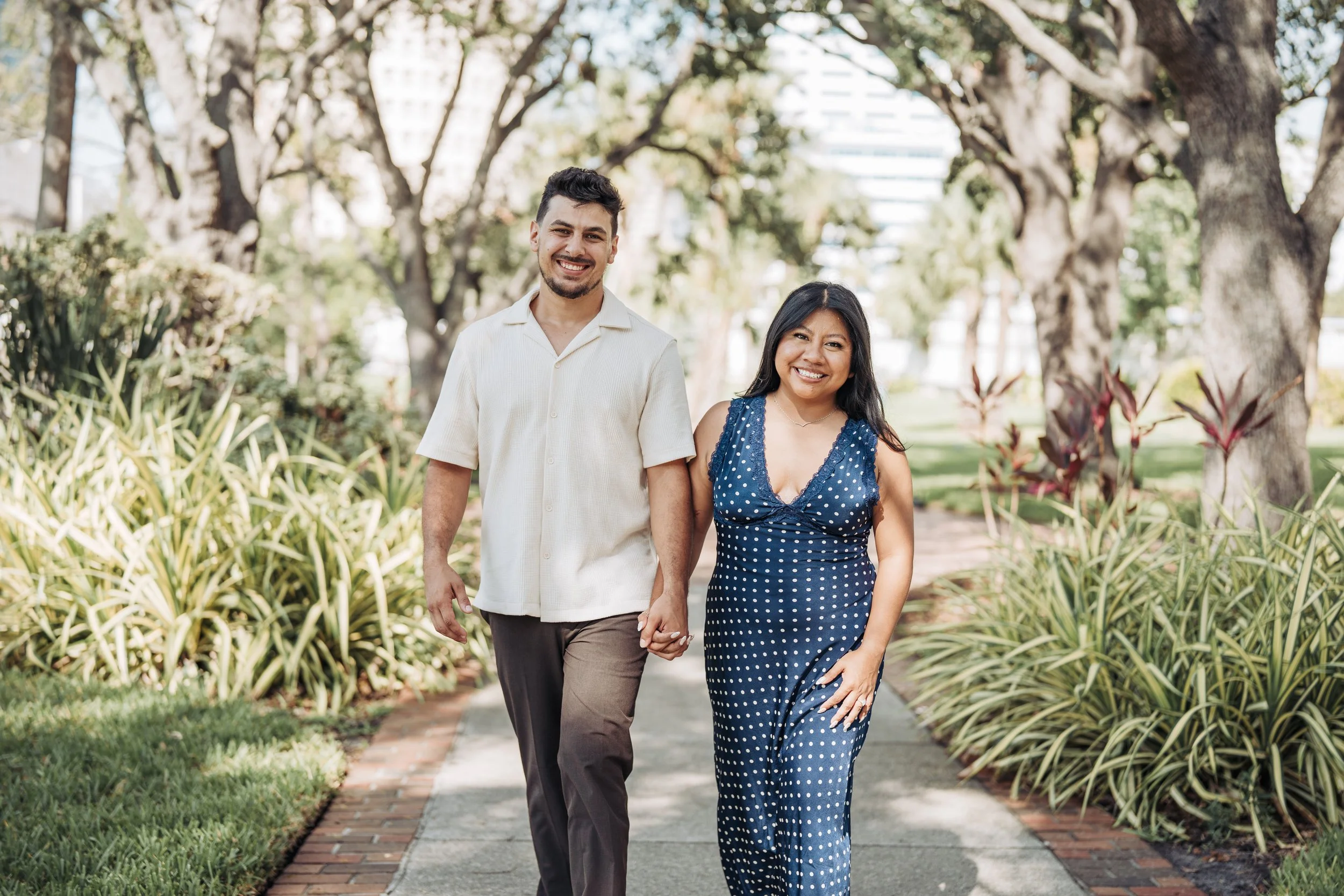 A smiling couple holding hands and walking together outdoors on a sunny day, surrounded by trees and greenery.