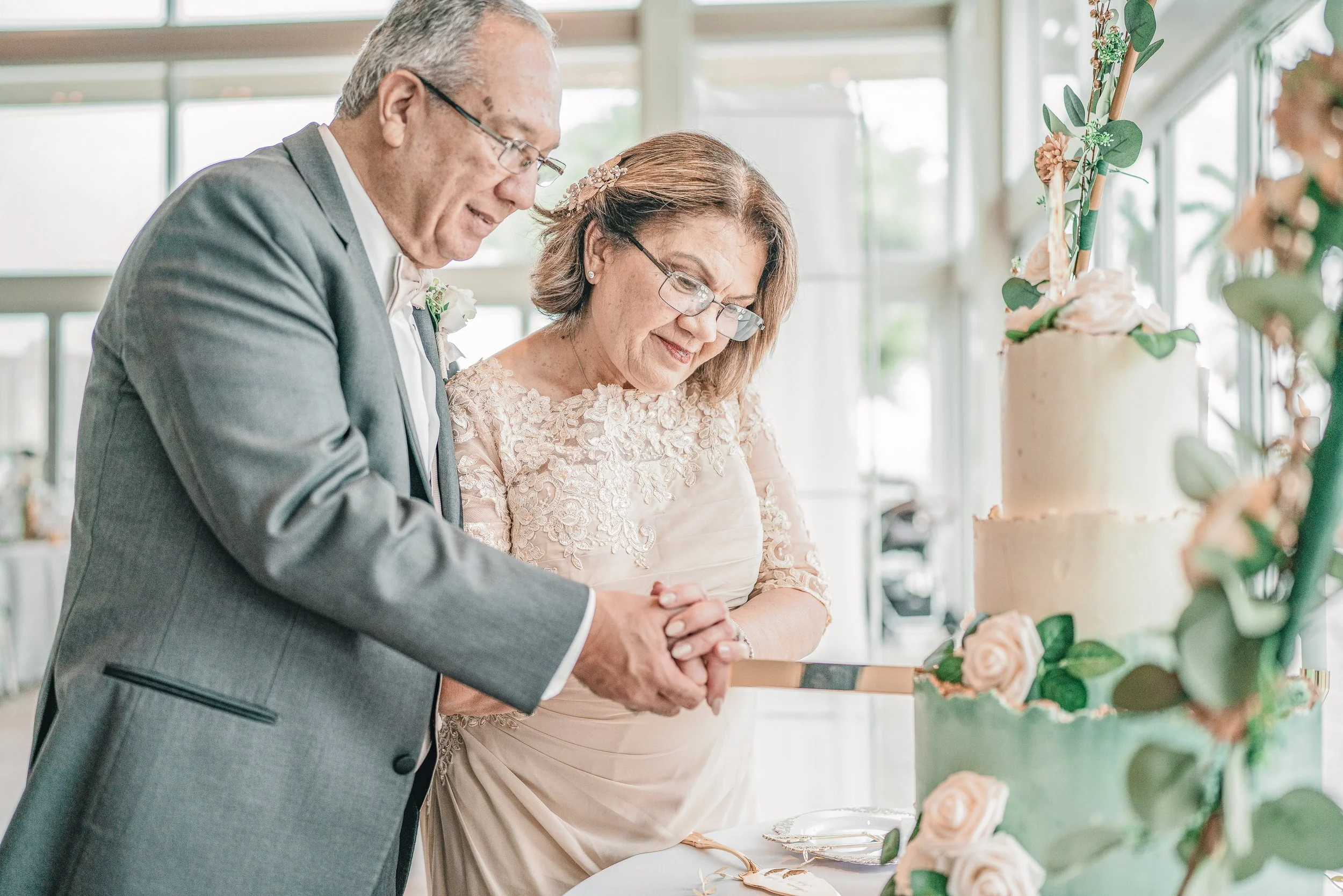An elderly man and woman, possibly a couple, are cutting a wedding cake together at a celebration. The man is wearing glasses and a gray suit, while the woman is dressed in a light-colored dress with lace details and a decorative hairpiece. They are 