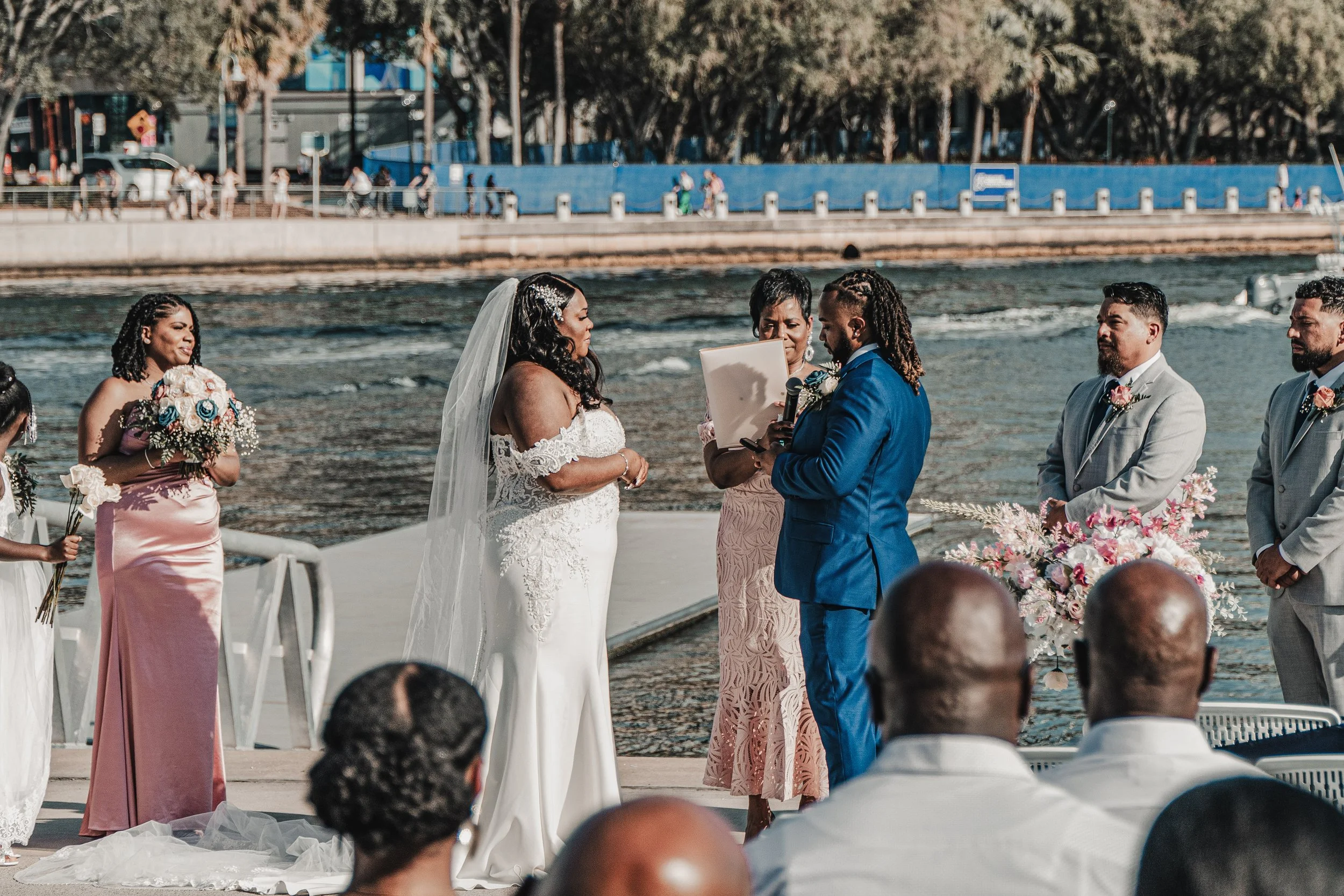 A wedding ceremony taking place outdoors by the water, with a bride and groom exchanging vows, surrounded by bridesmaids and groomsmen.