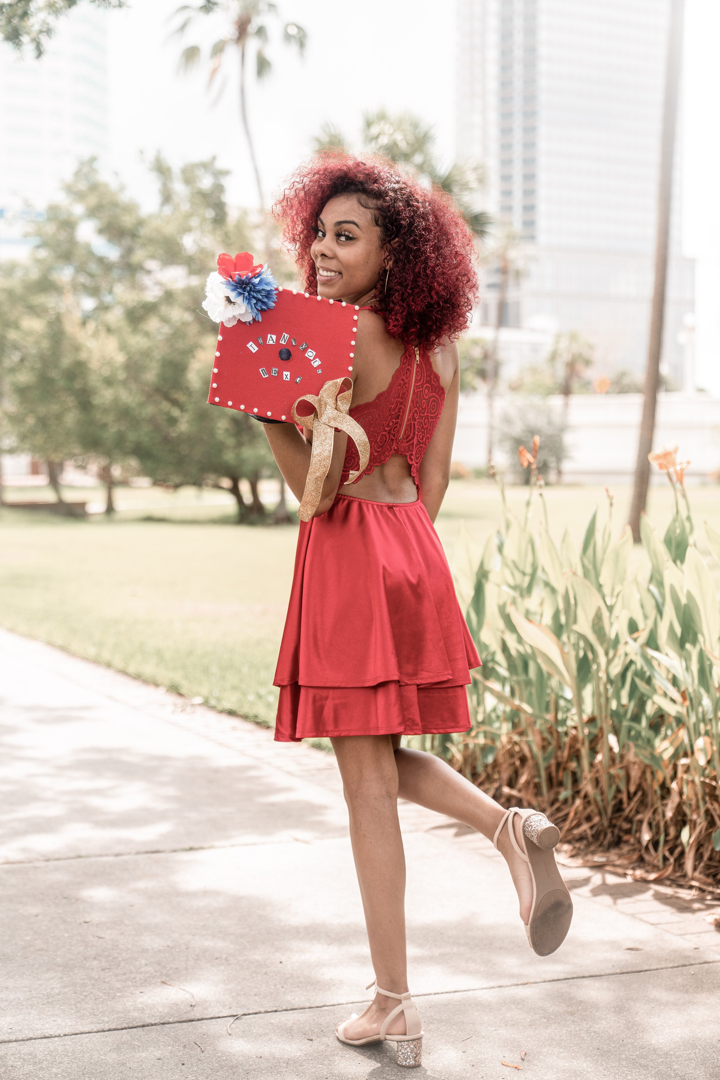 Young woman in red dress with curly red hair posing outdoors, holding a decorated graduation cap with the words "Thank You IELTS" on it, surrounded by greenery and tall buildings in the background.
