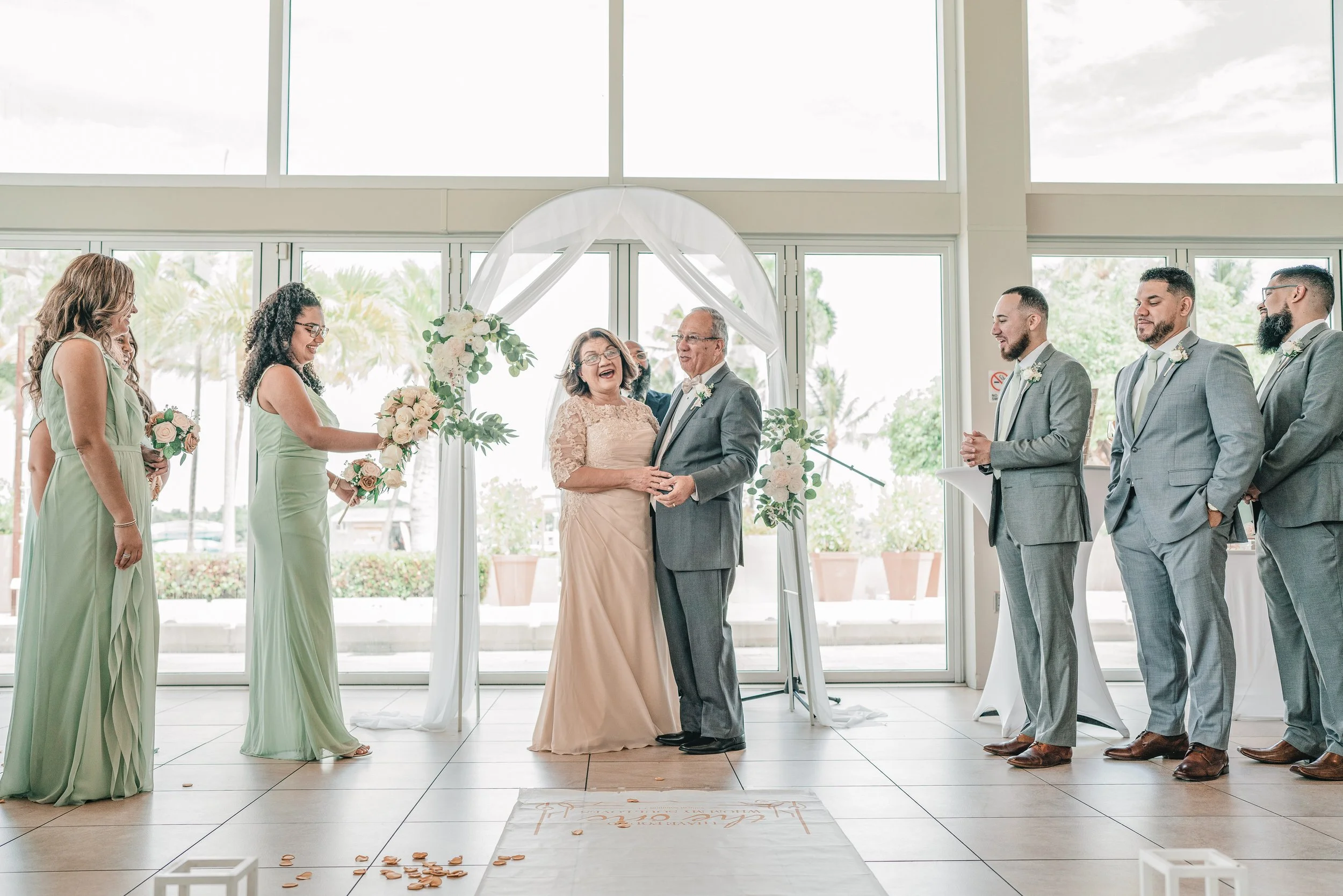 Wedding ceremony with bride, groom, bridesmaids, and groomsmen standing in a bright indoor space with large windows, floral decorations, and an arch behind the couple.