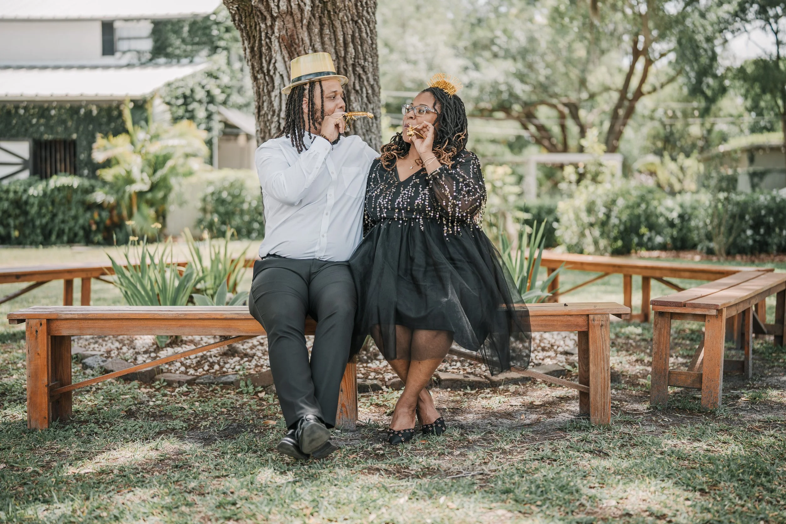 A couple sitting on a wooden park bench, wearing party hats and celebration accessories, blowing party horns at a birthday or celebration event outdoors.