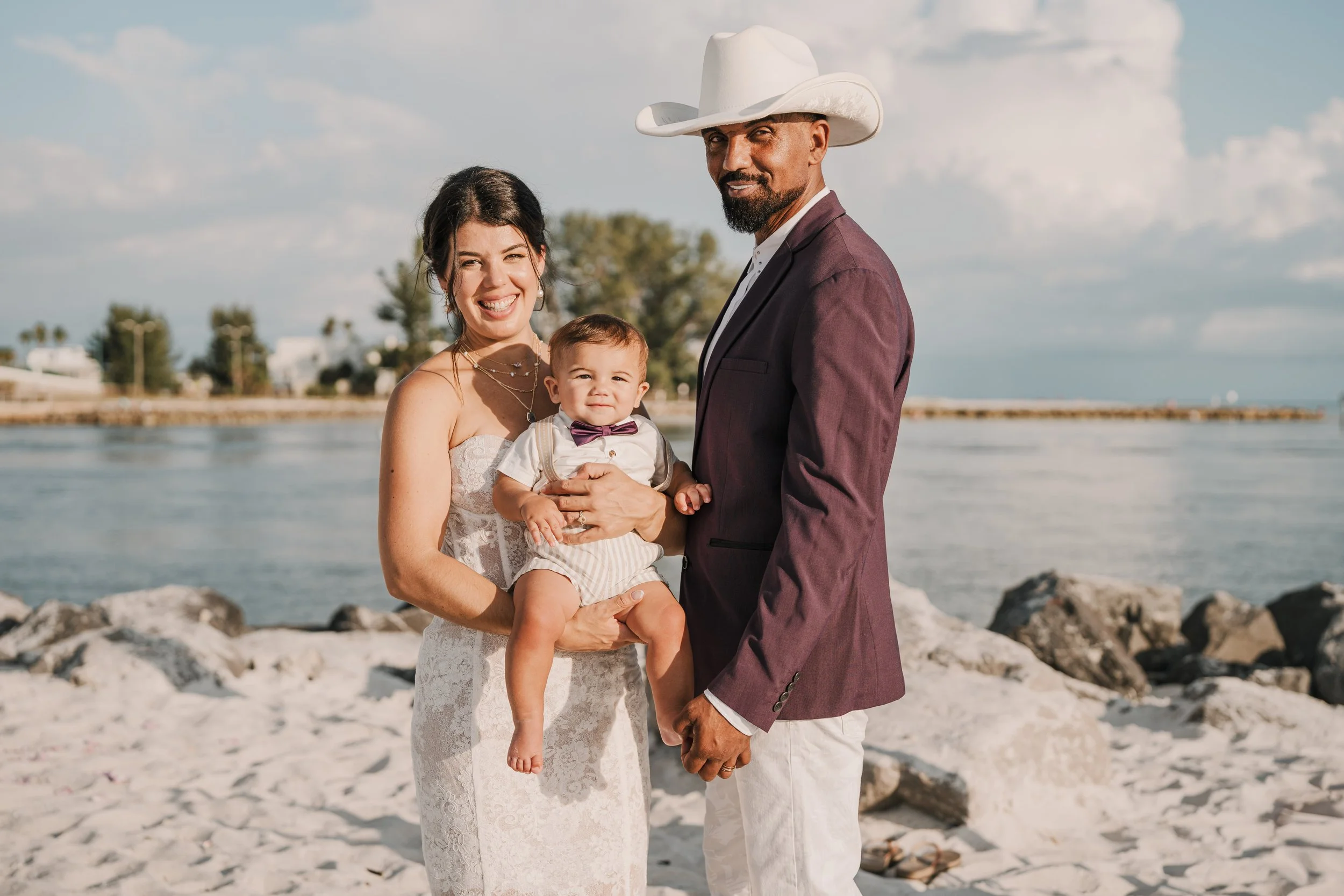 A family of three at the beach, with a woman in a white lace dress holding a baby boy dressed in a white shirt with suspenders and a bow tie, and a man in a maroon suit and white cowboy hat standing beside them.