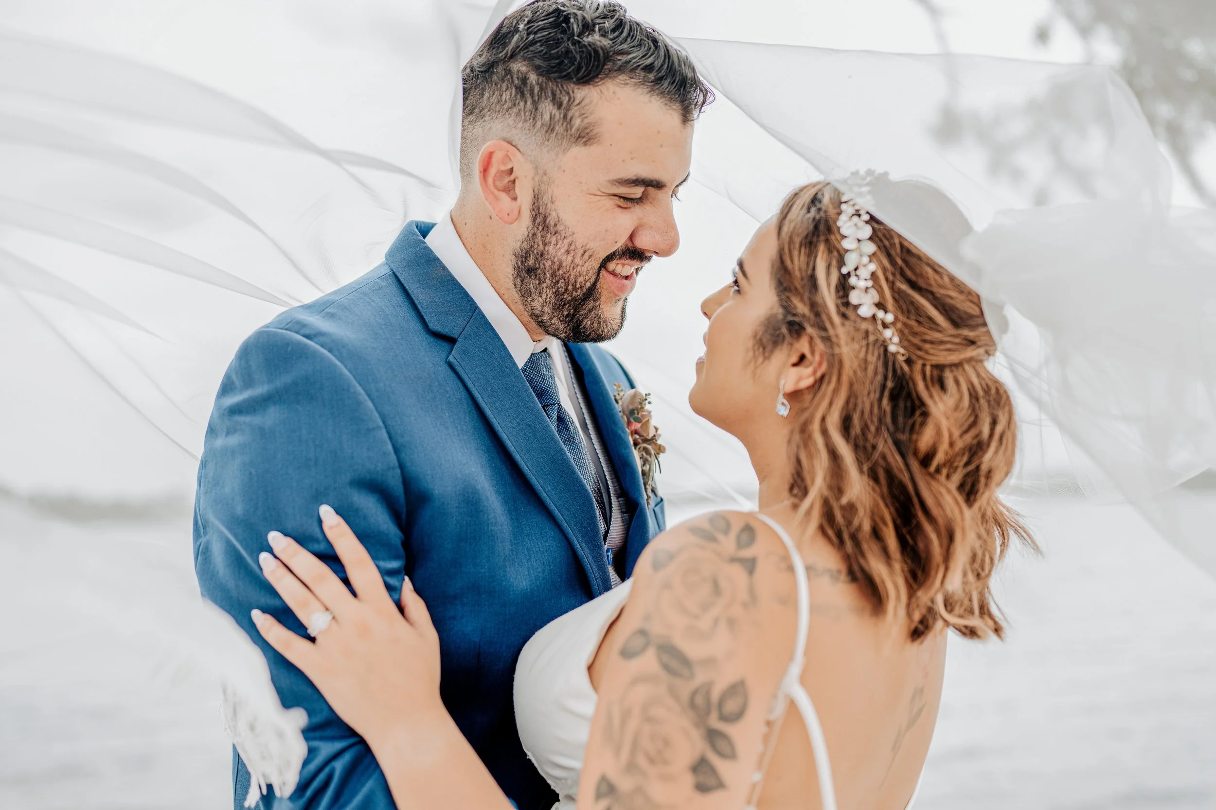 A couple, a man in a blue suit and a woman with tattoos, are standing close under a white fabric, looking at each other with smiles on their faces.