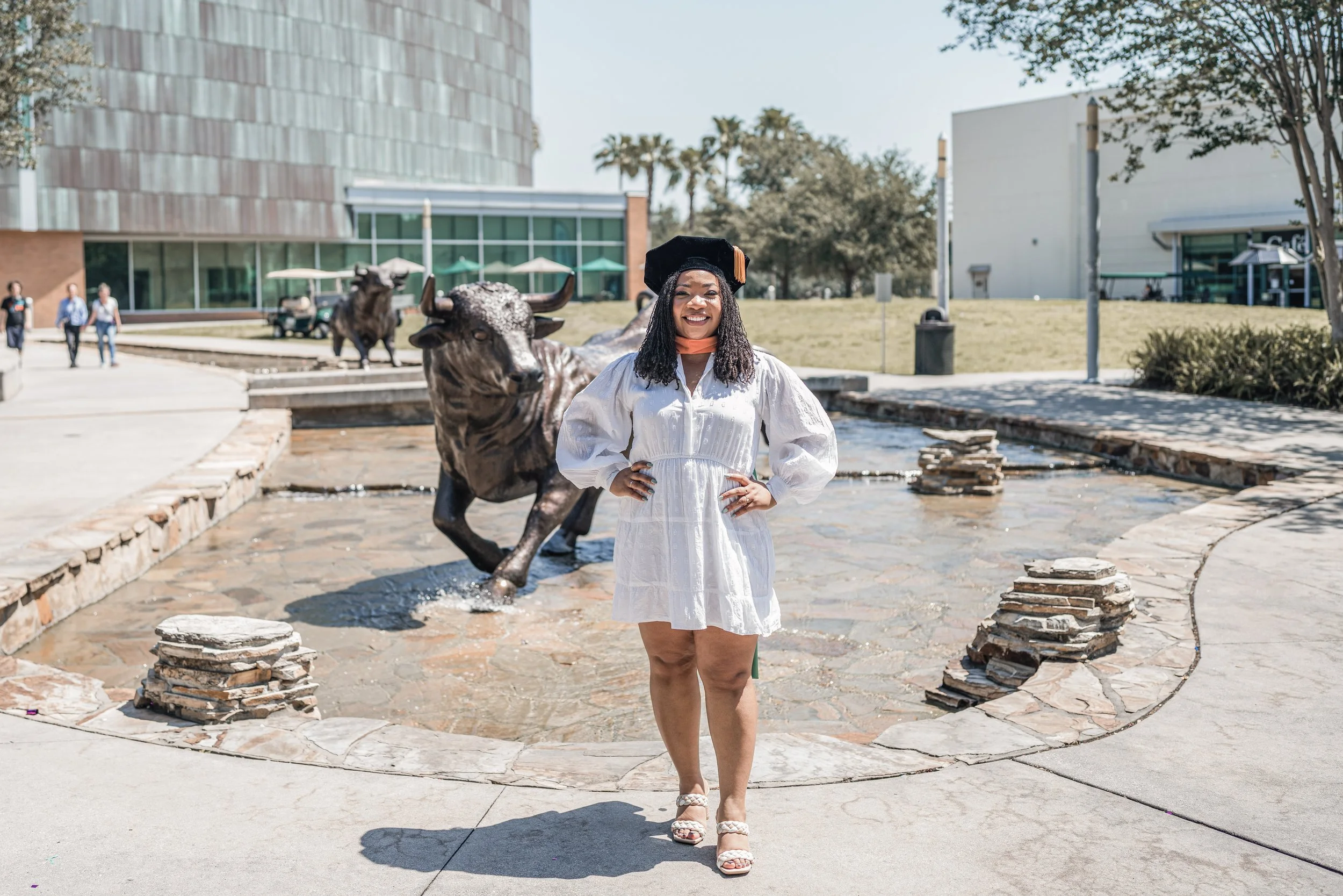 A woman in a white dress and black graduation cap stands in front of a fountain with a bronze statue of a buffalo. She is smiling and has her hands on her hips. There are trees, buildings, and people in the background on a sunny day.