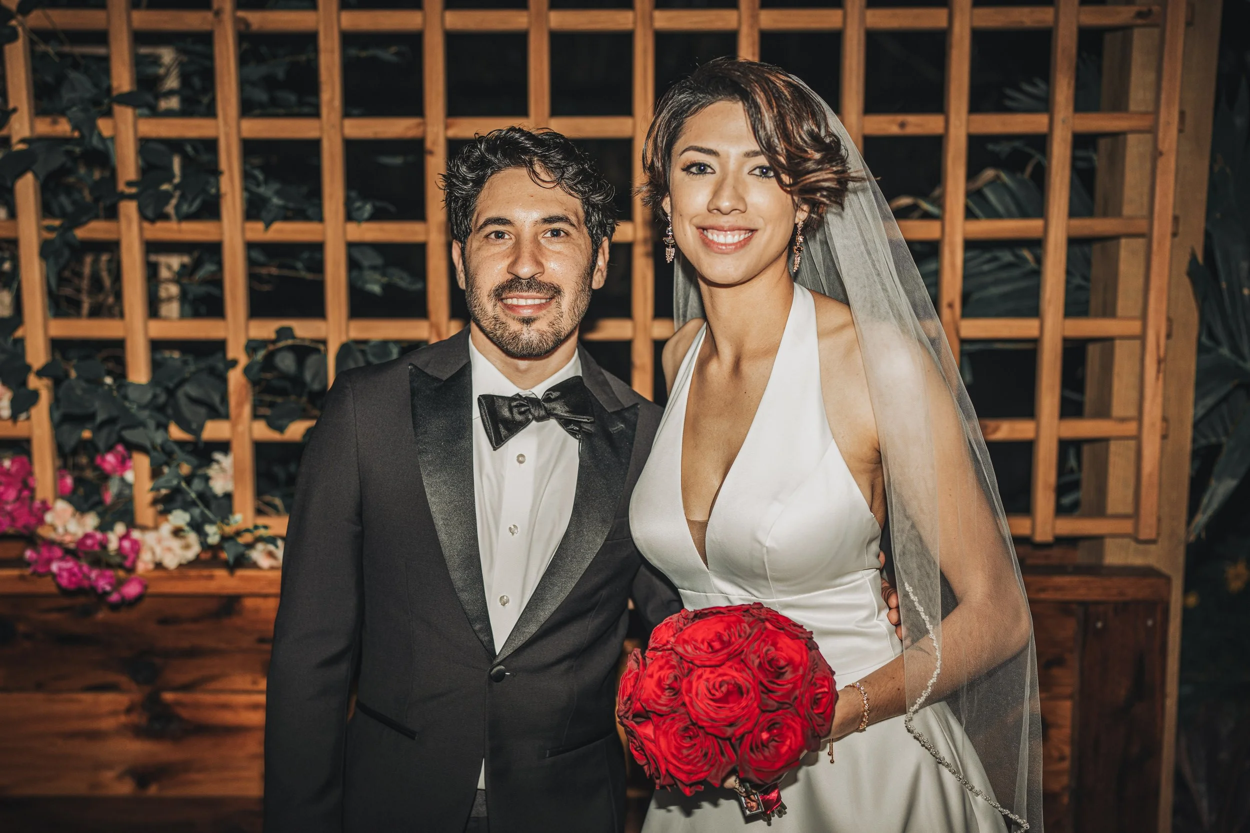 A smiling bride and groom at their wedding, standing in front of a wooden lattice backdrop with greenery and flowers.