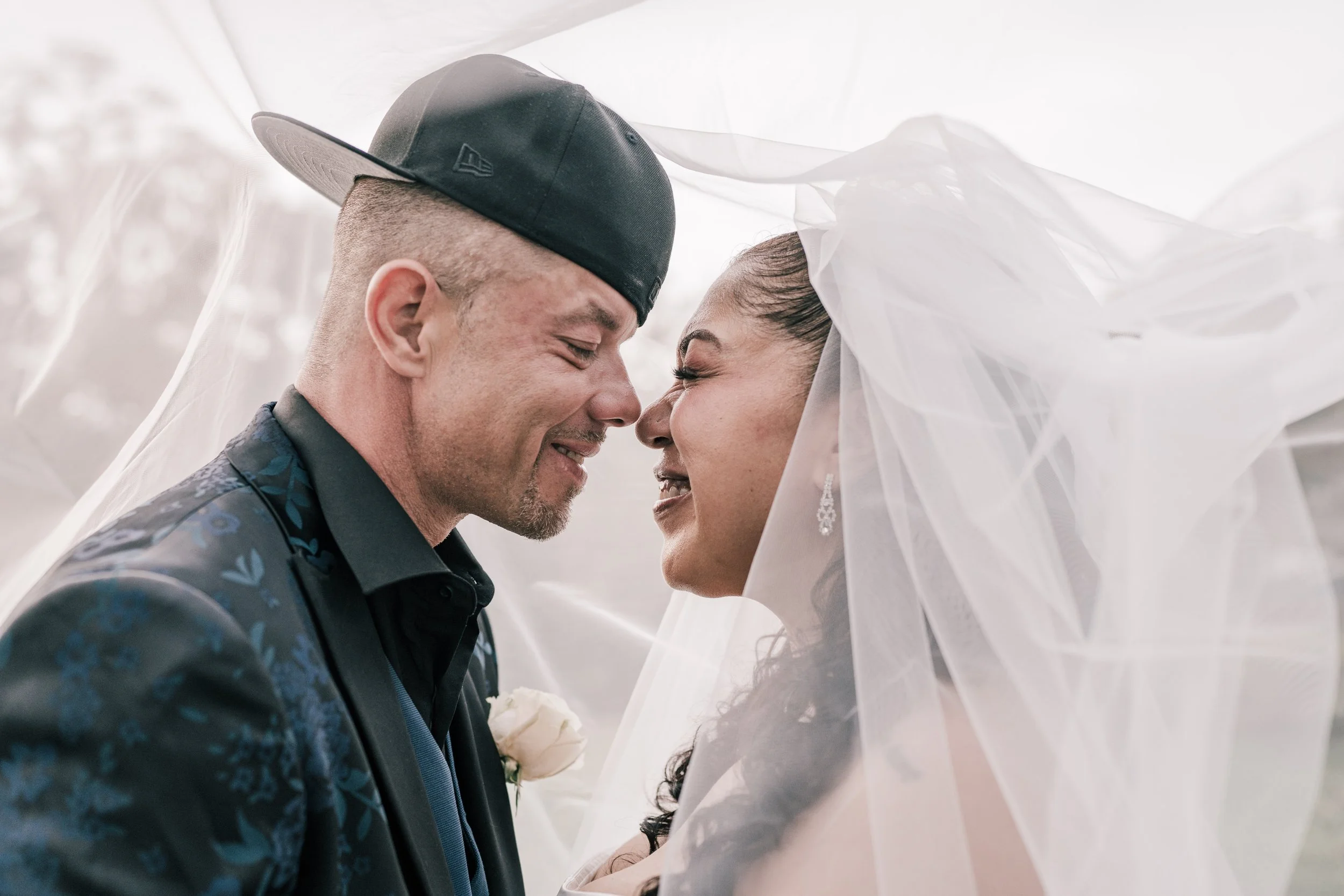 A man and woman, dressed in wedding attire, are smiling and about to kiss under a white fabric canopy, celebrating their wedding.