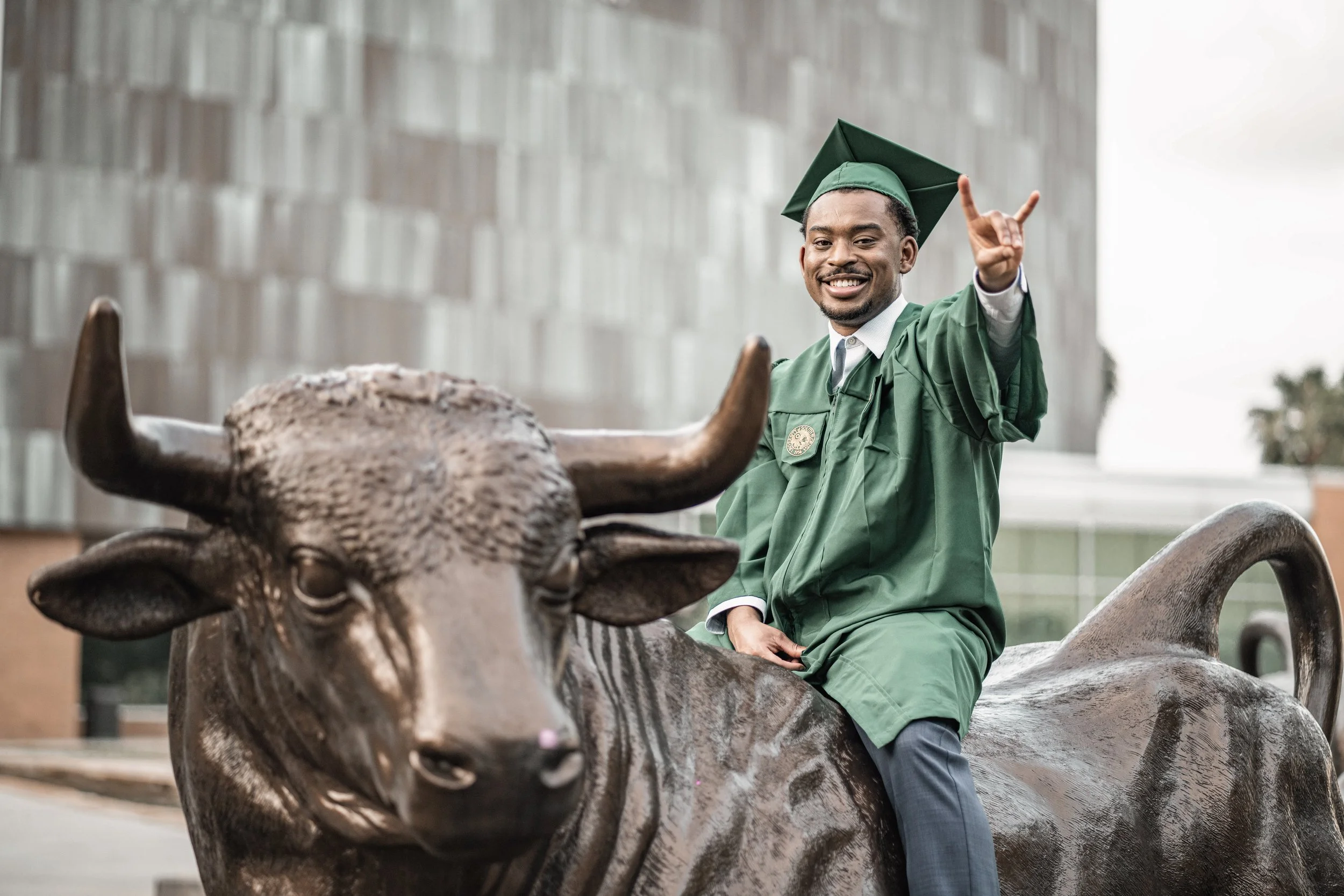 A young man in a green graduation gown and cap, smiling and making a hand gesture, sitting on a large animal sculpture of a buffalo or bison.