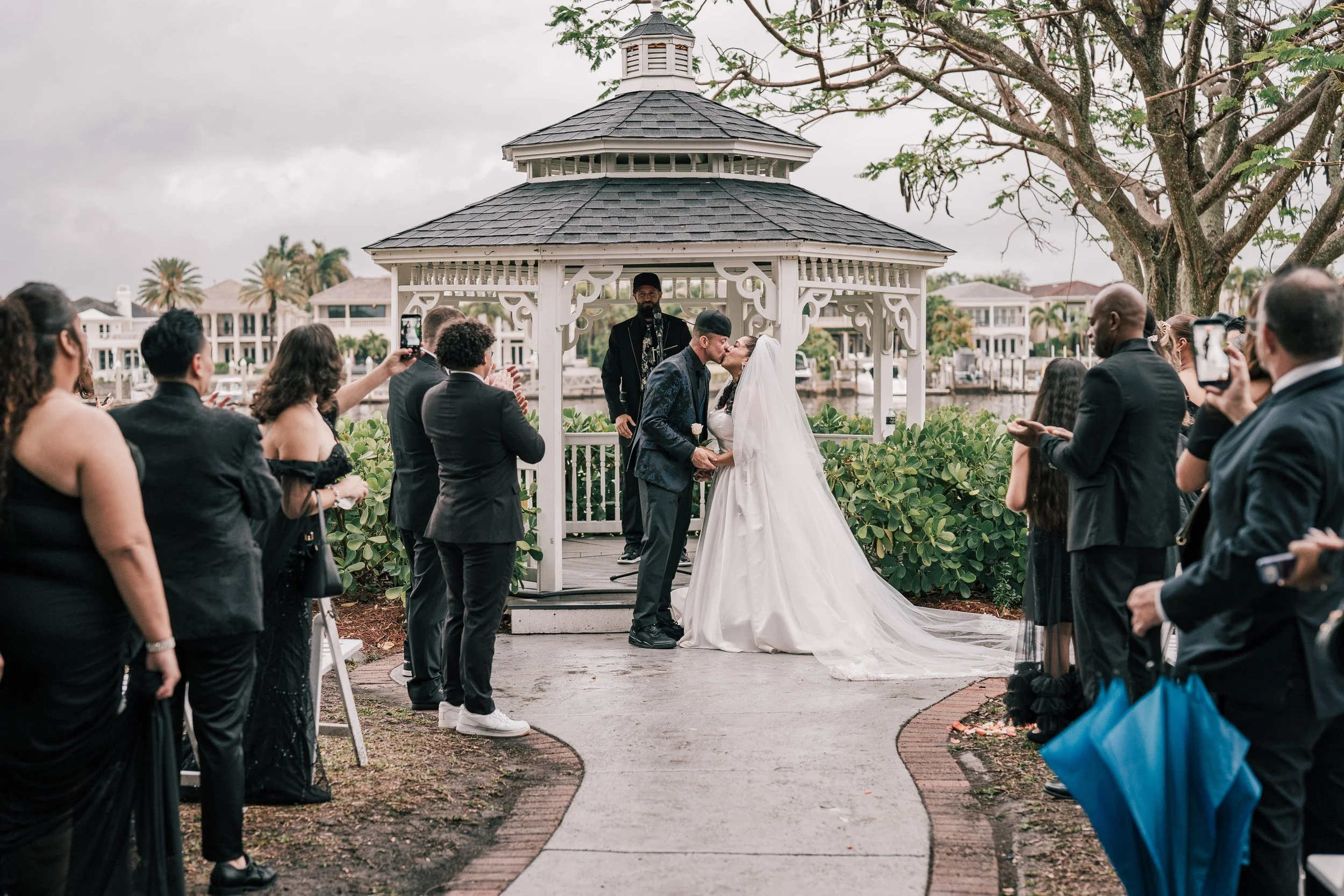 A couple shares a kiss during their outdoor wedding ceremony under a white gazebo, surrounded by family and friends.
