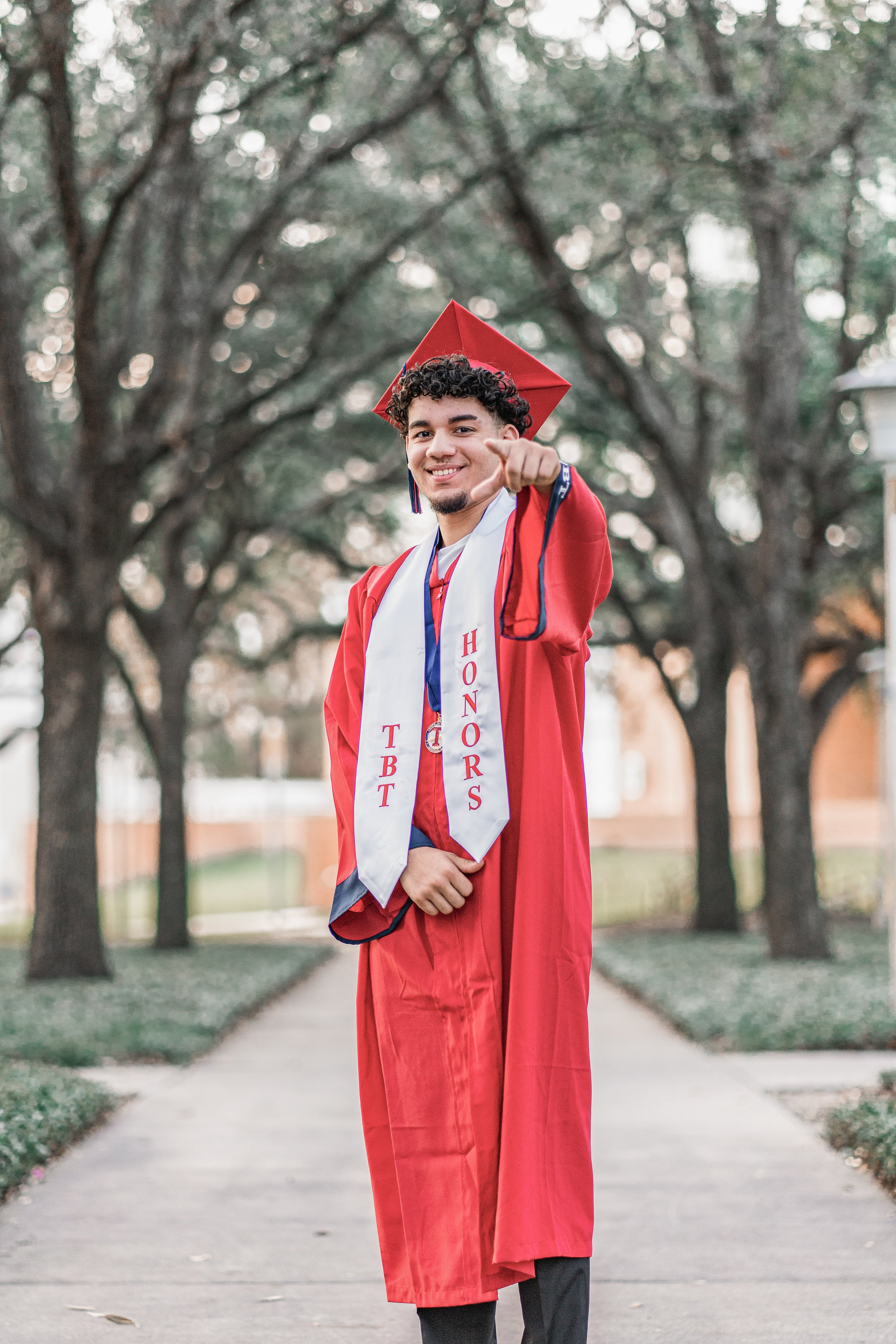 A young man in a red graduation gown and cap is standing outdoors in front of tree-lined walkways and smiling, pointing at the camera.