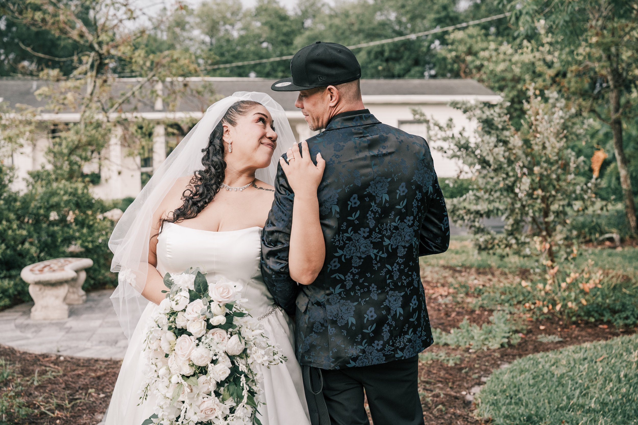 A bride and groom share a romantic moment outdoors, standing close with the bride looking up at the groom, holding a bouquet of white roses, surrounded by trees and fallen leaves.