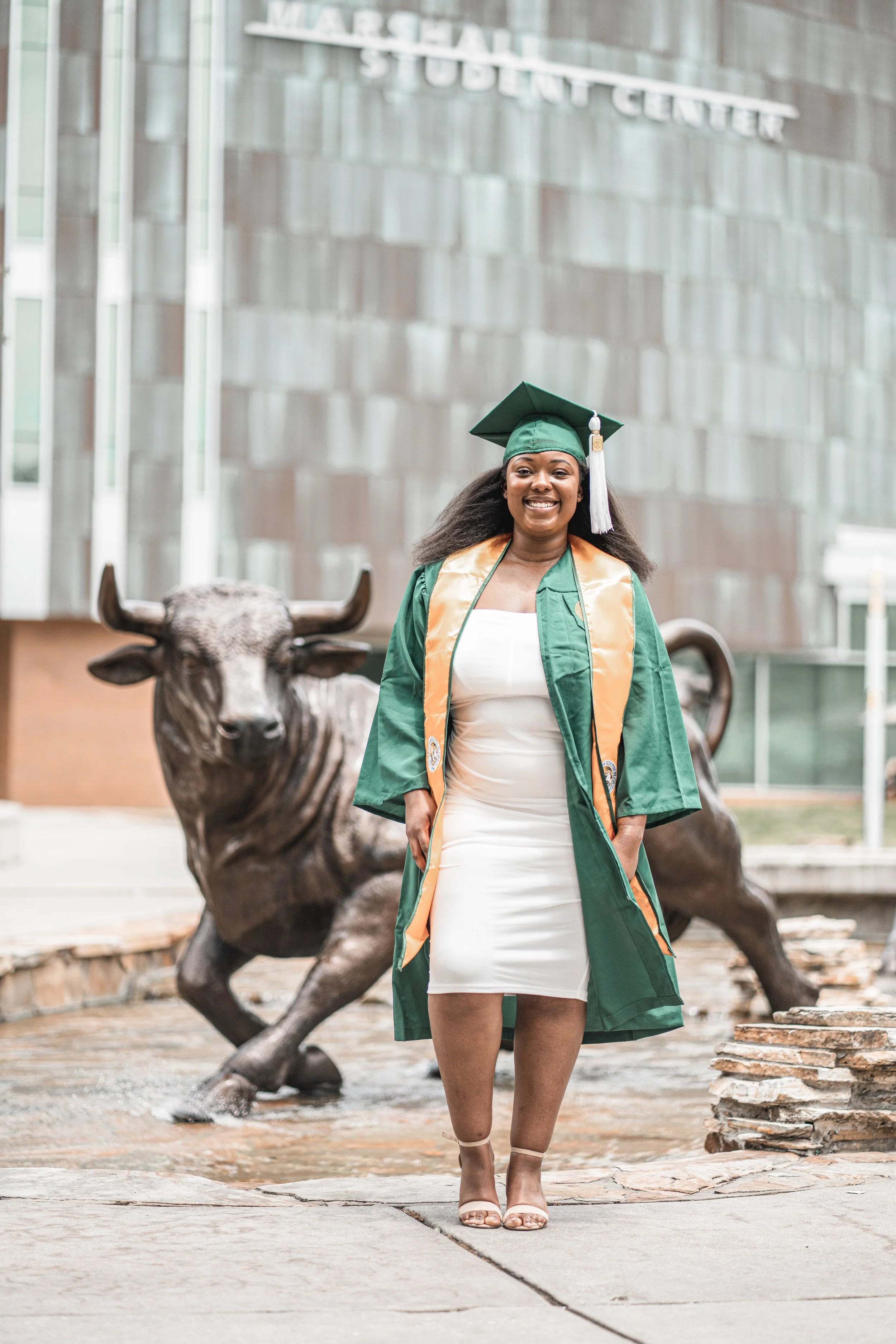 A woman in a green graduation gown and cap standing in front of a bronze statue of a bull at a university campus.