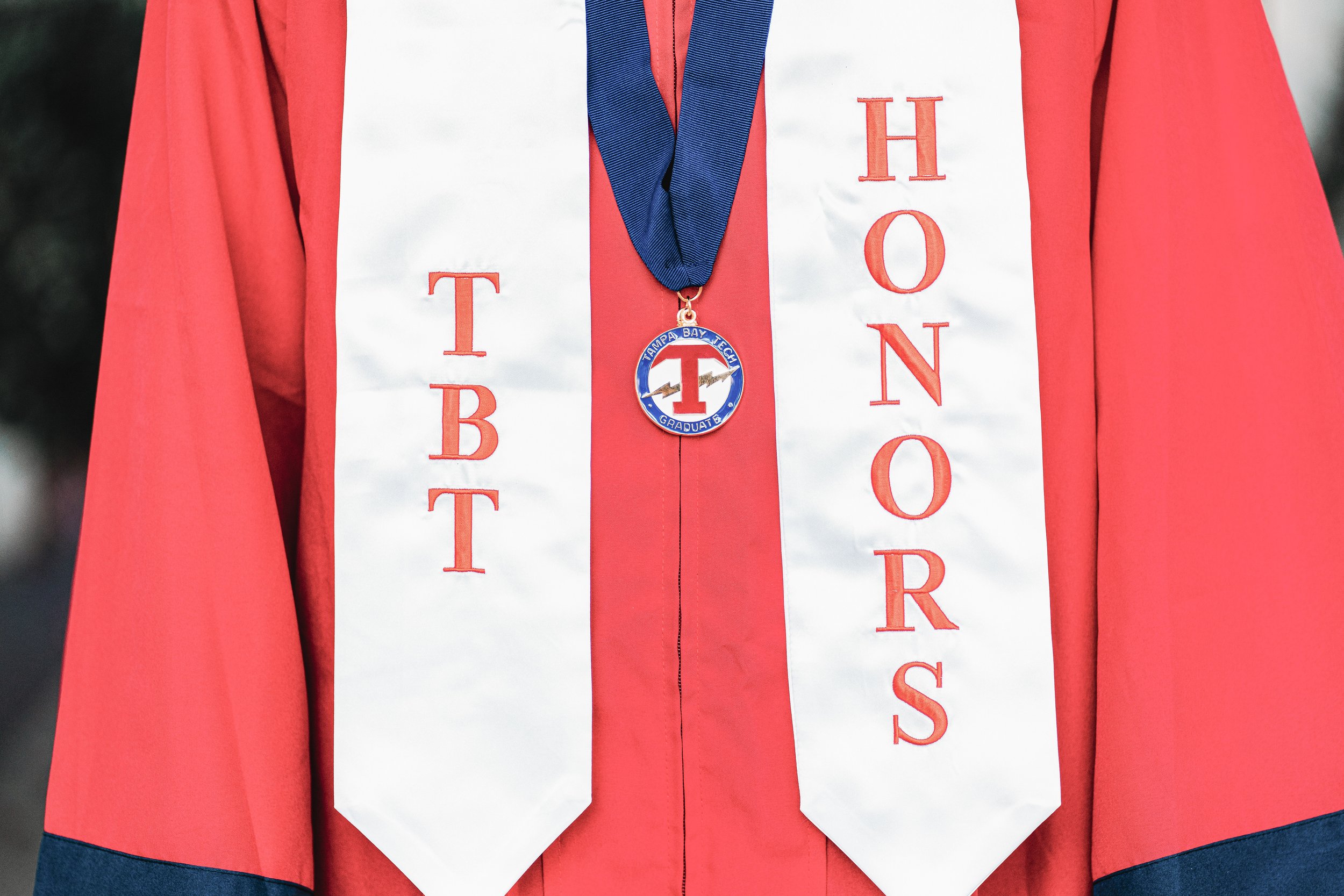 A person wearing a red graduation gown and a white stole with red embroidered letters, with a navy blue graduation medal hanging around their neck.