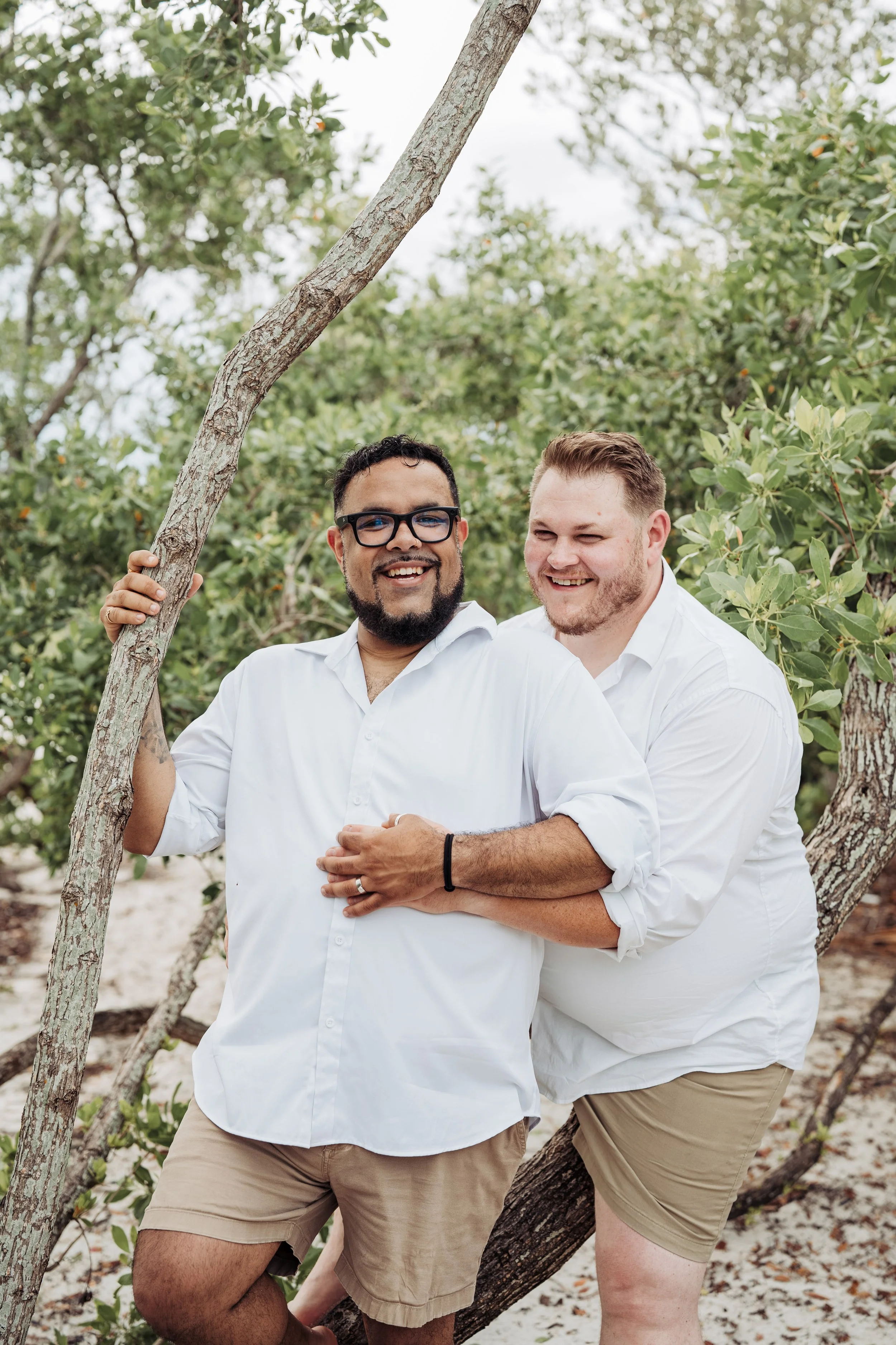 Two men standing in front of trees and greenery, smiling and embracing each other outdoors.
