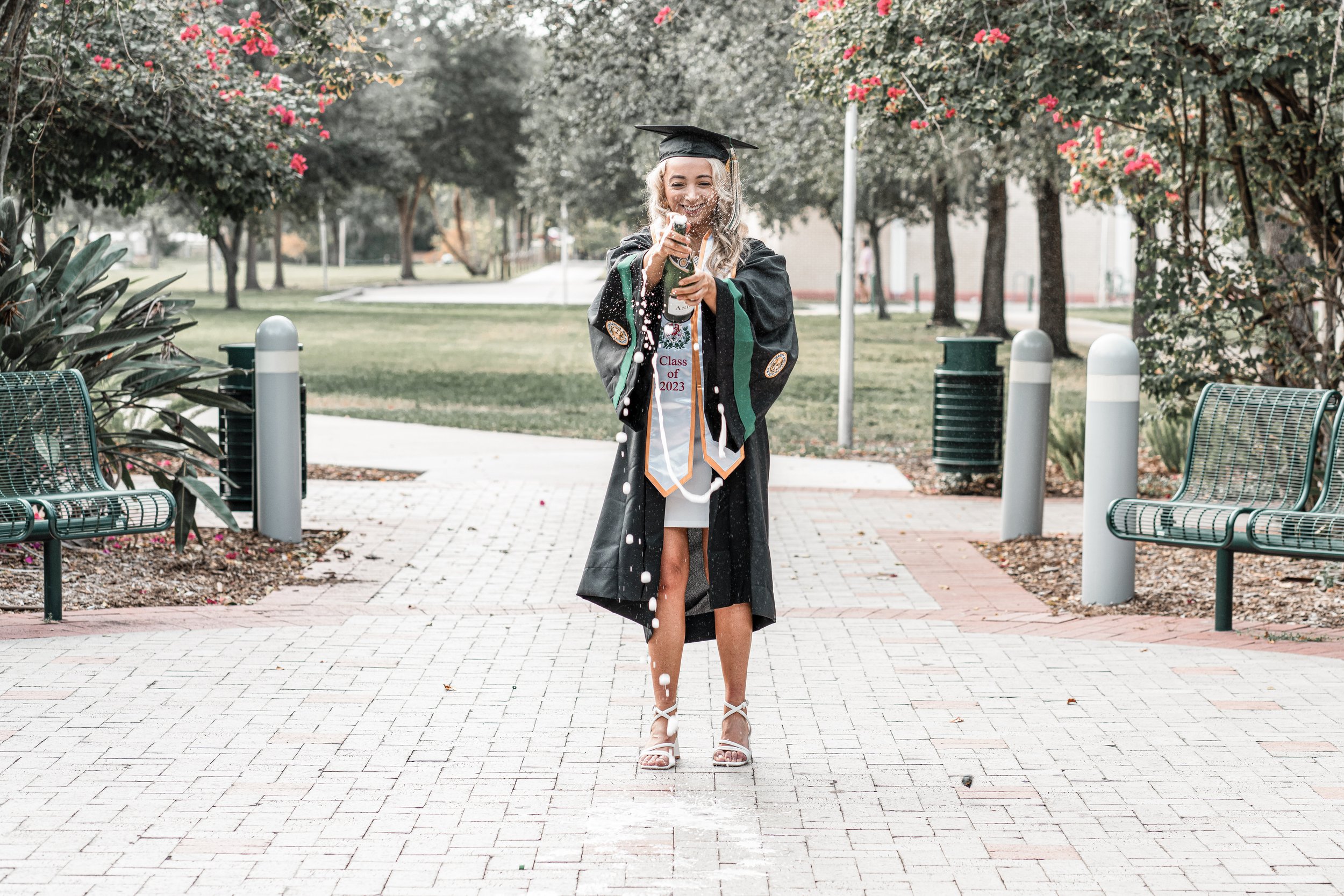 A woman in a graduation cap and gown celebrating outdoors, smiling and spraying champagne.