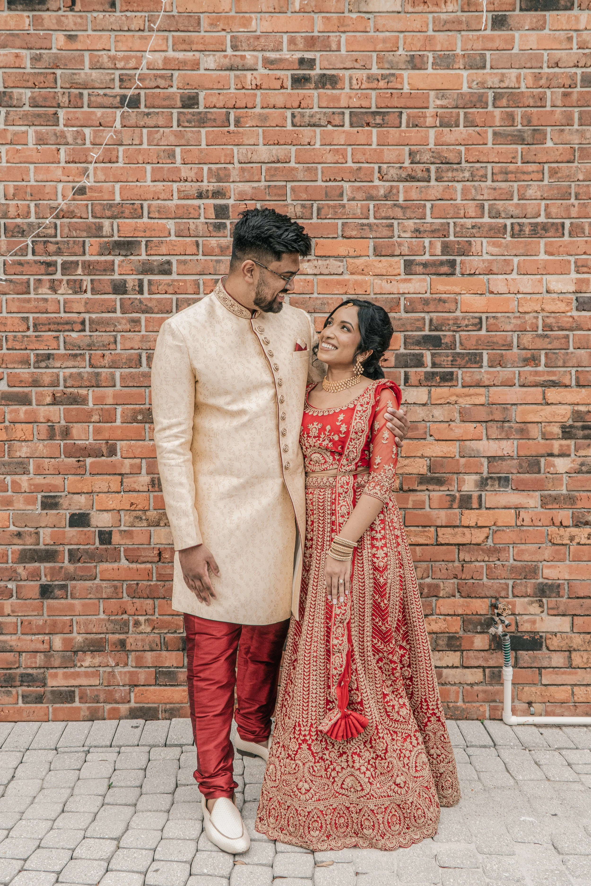 A couple dressed in traditional Indian attire standing against a brick wall. The man wears a cream-colored embroidered sherwani with red pants, and the woman wears a red and gold lehenga with intricate embroidery, gold jewelry, and is looking up at t