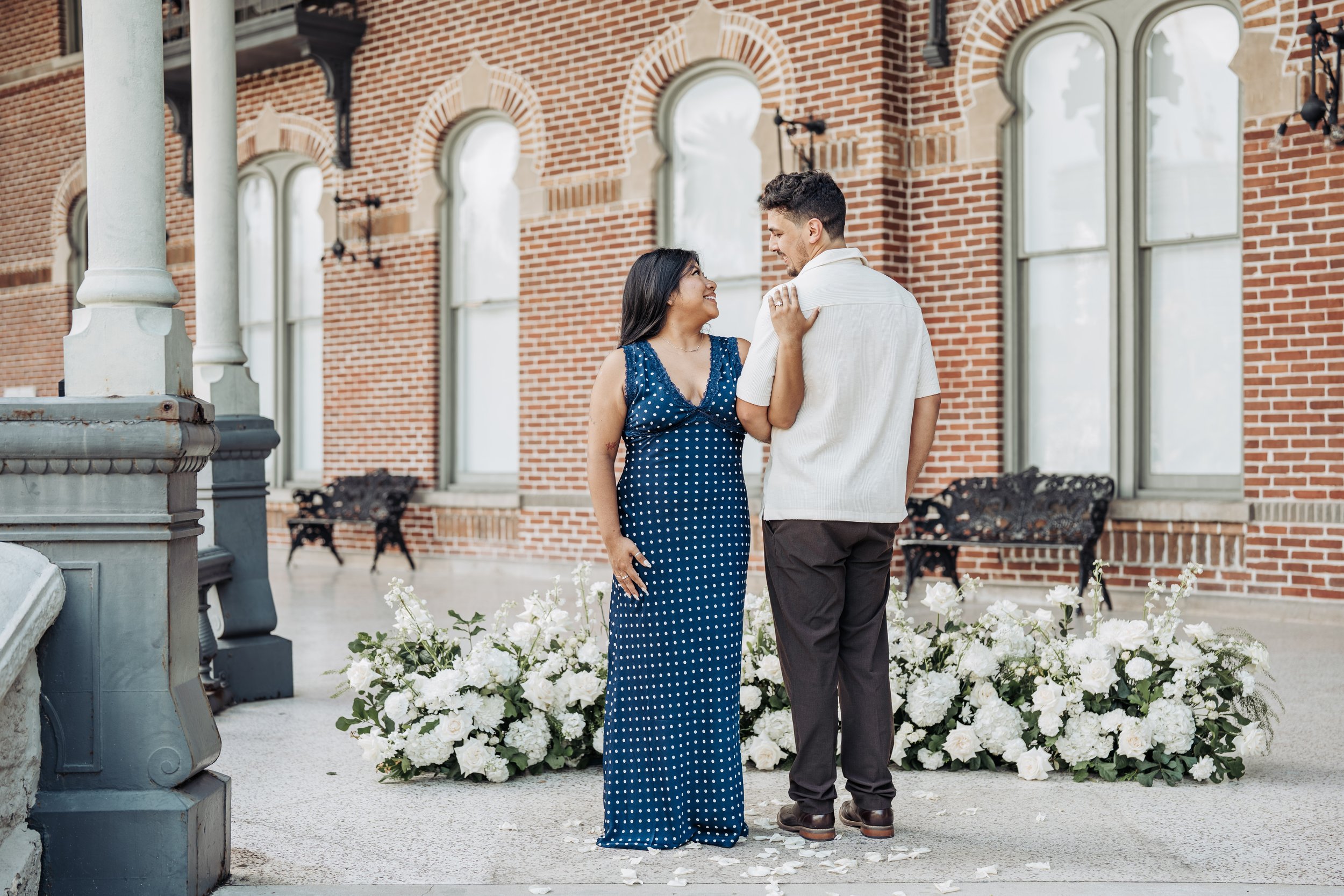 A couple stands close together outdoors, smiling and looking into each other's eyes, with a floral arrangement and brick building in the background.