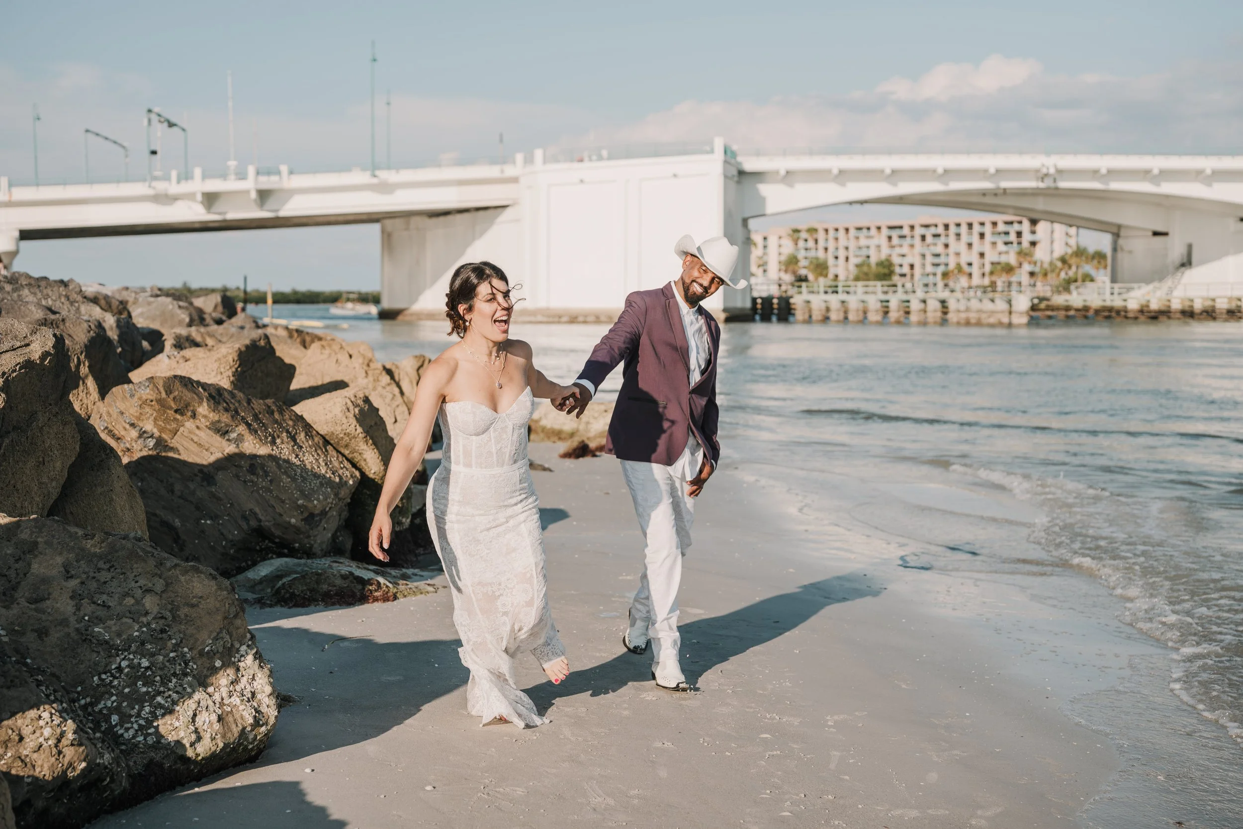 A bride and groom holding hands and walking on the beach near rocks, with a bridge and buildings in the background, under a partly cloudy sky.